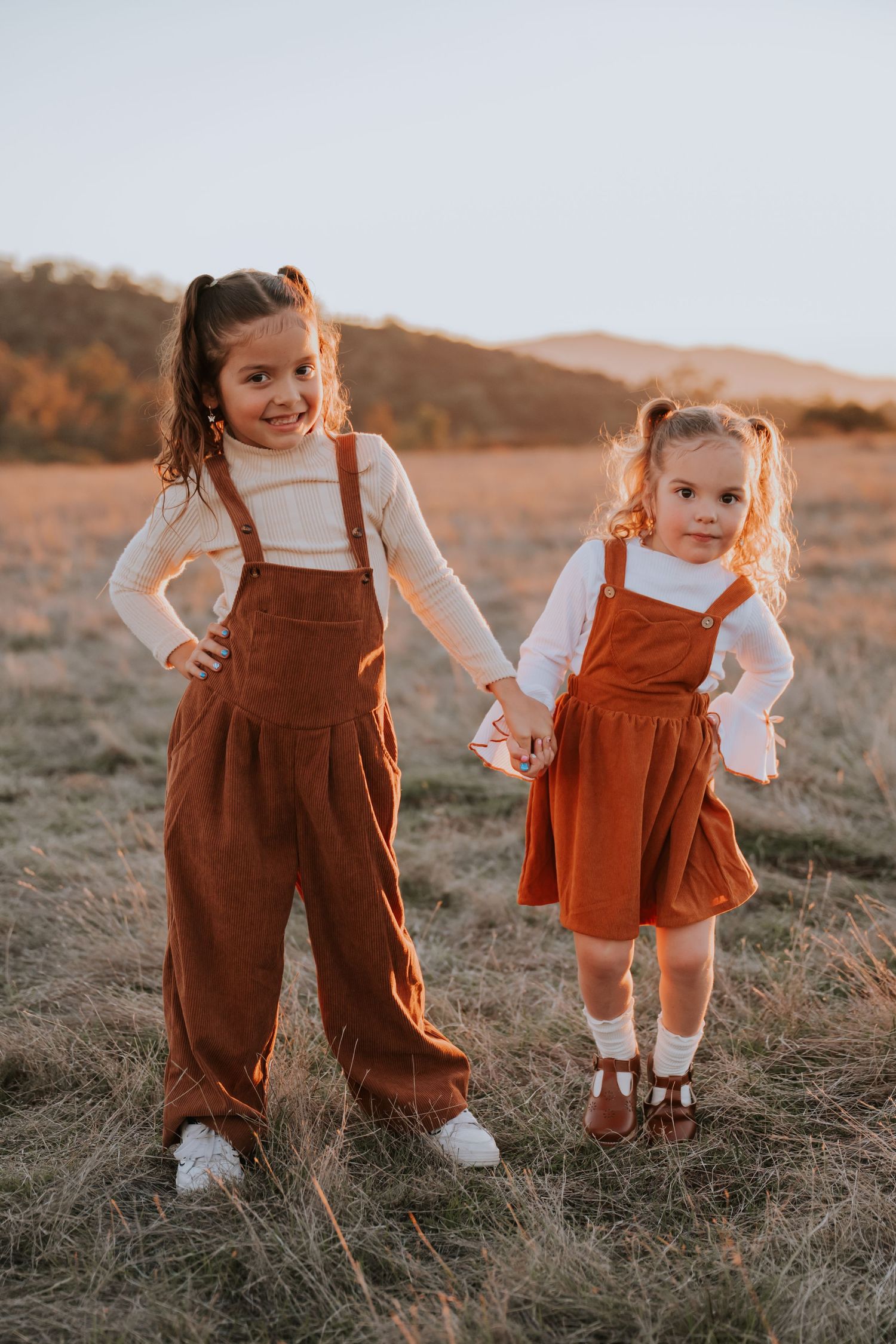 Two young girls in matching rust-colored overalls hold hands in a golden field at sunset.