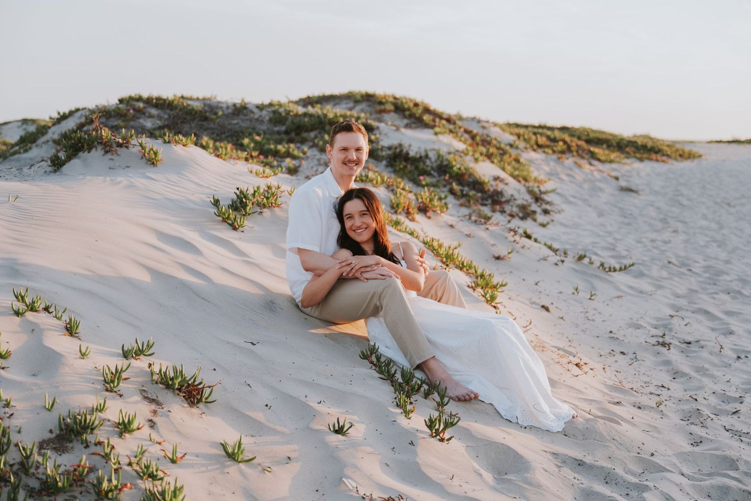Couple sitting on white sand dunes at sunset, woman in white dress, man in white shirt, smiling together outdoors.