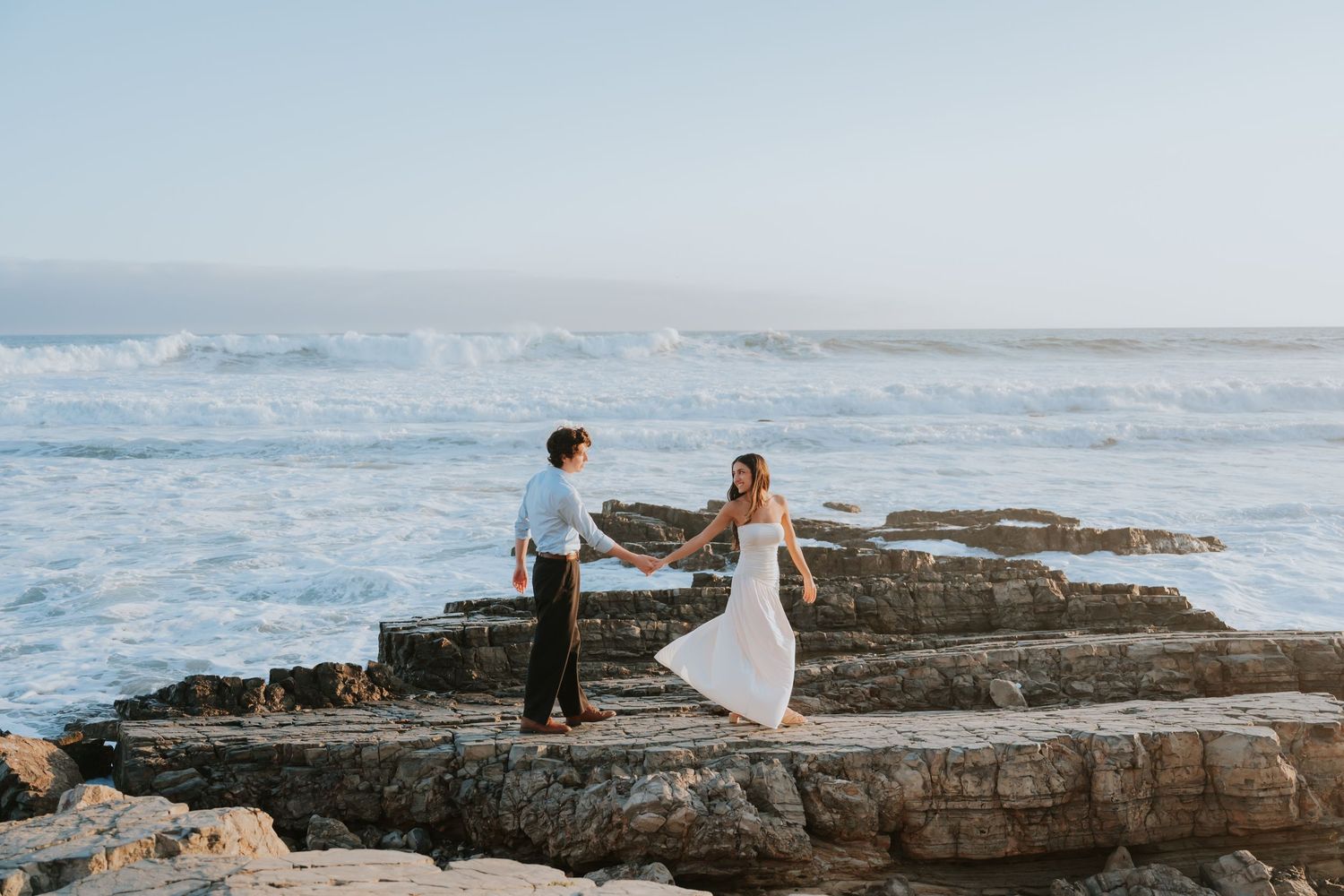 Couple holds hands on rocky coastal cliffs with crashing ocean waves behind them during a romantic engagement photo session.