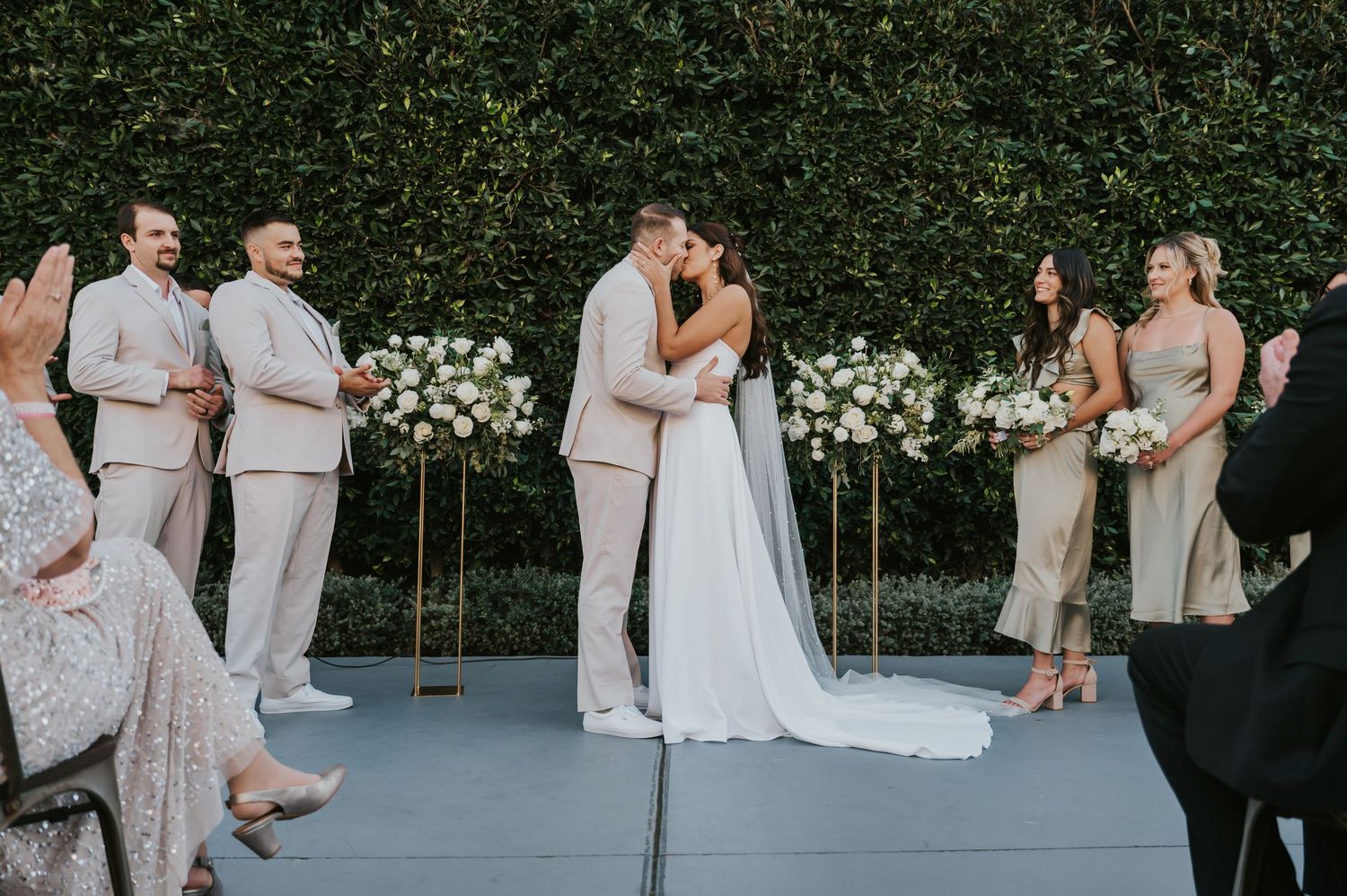 Wedding party in formal attire poses for candid photos in front of a lush green hedge wall with white flowers.