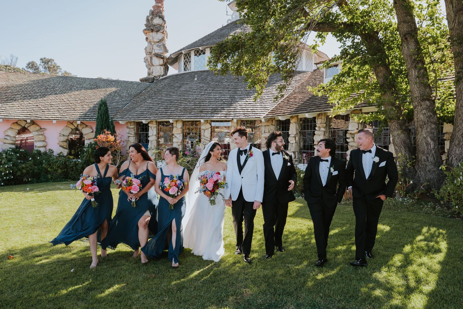 Wedding party in formal attire poses together on a grassy lawn with trees in the background.