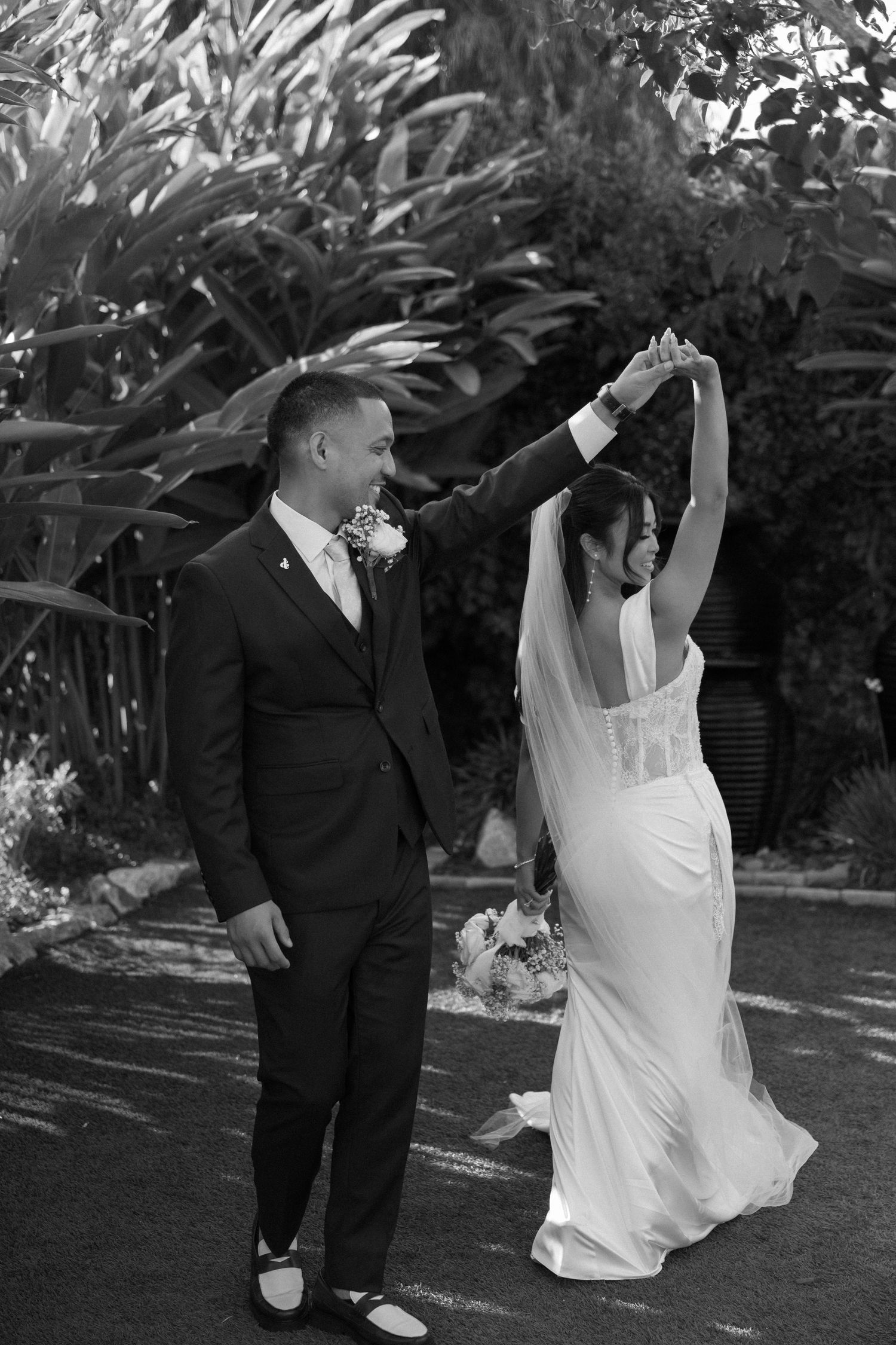 A black and white photo of newlyweds dancing in a garden, with the bride twirling in her elegant dress.