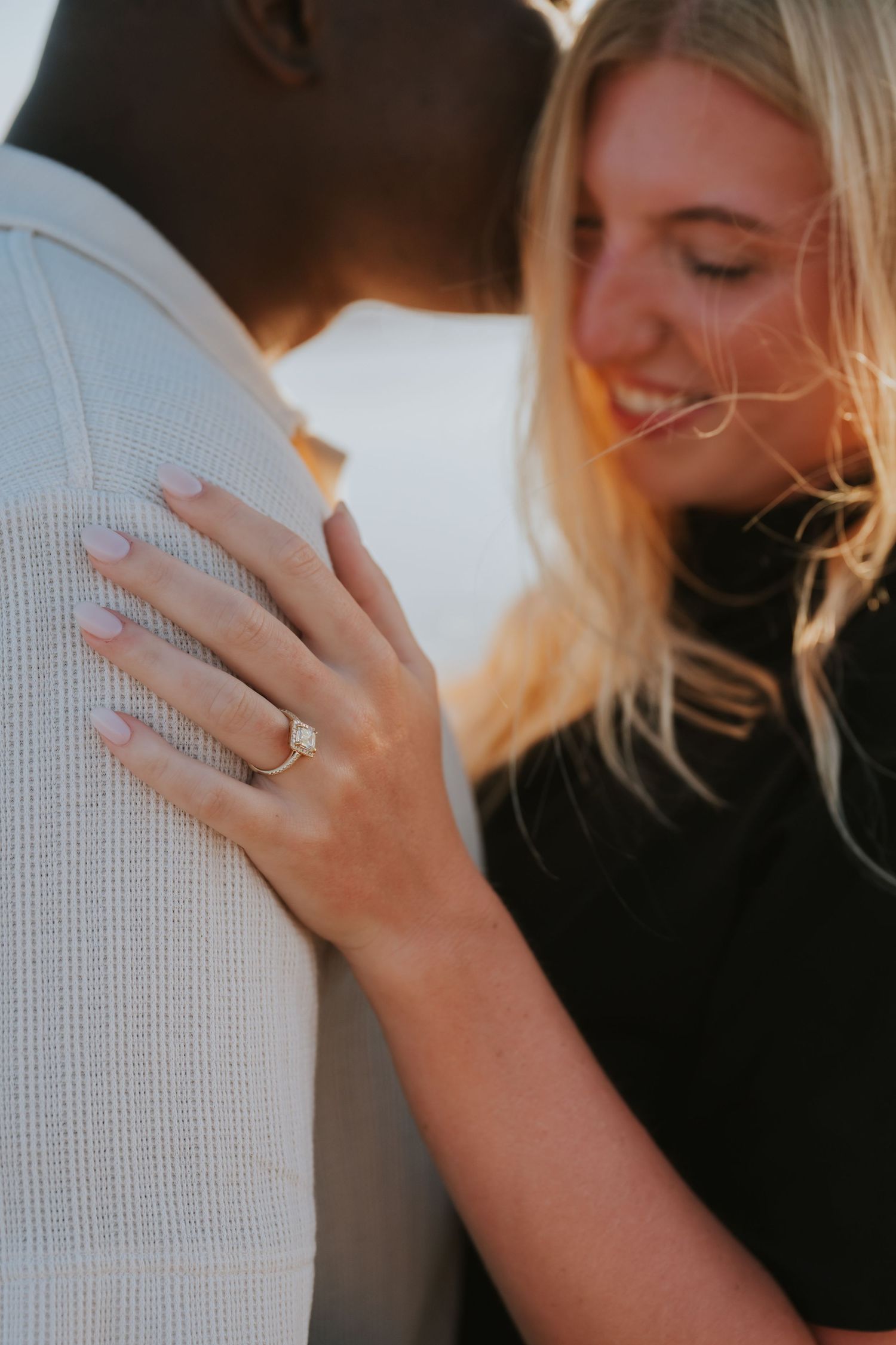 Smiling blonde woman rests hand with diamond engagement ring on man's shoulder in warm golden light.