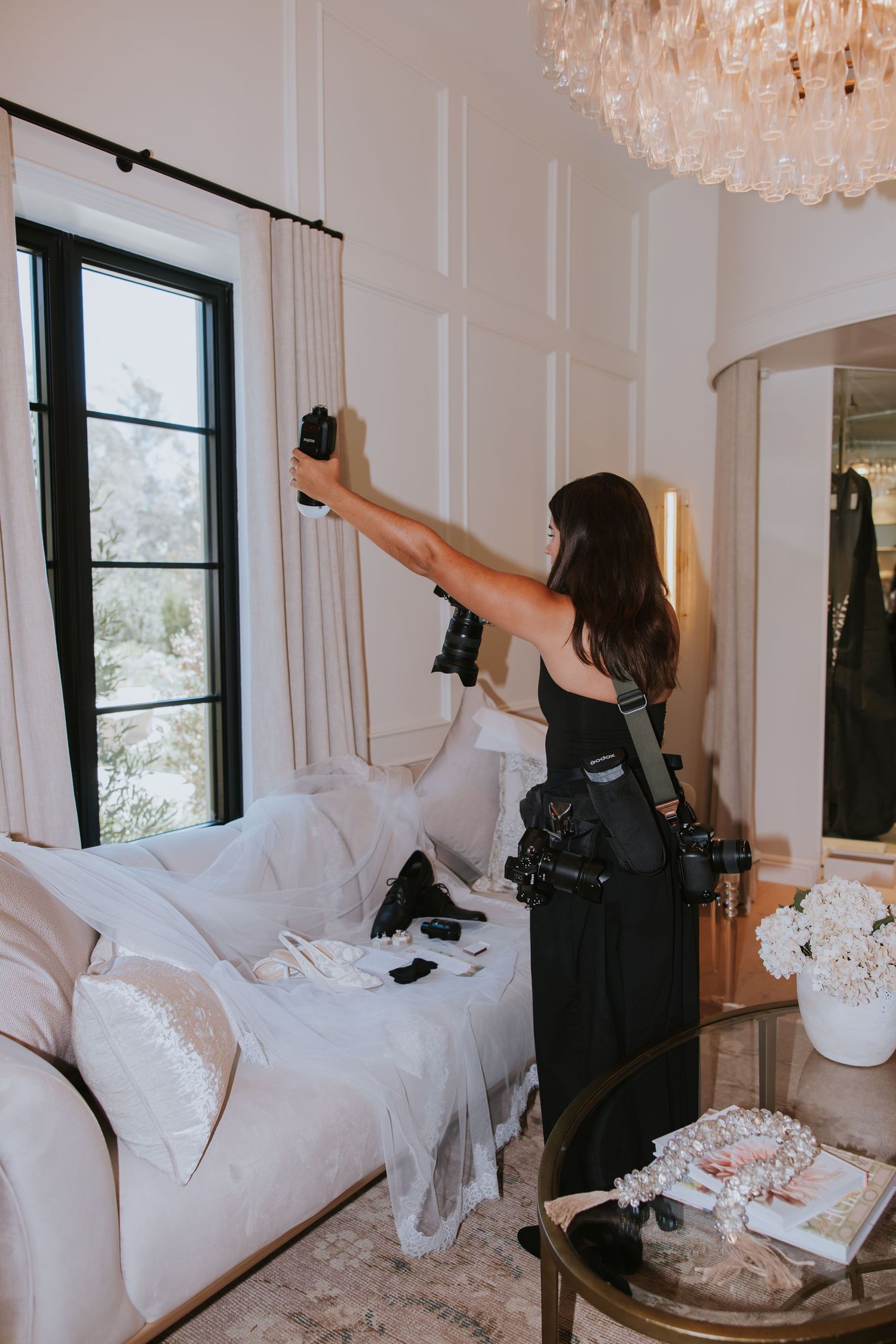 Photographer holding camera flash in elegant white bedroom with chandelier, sofa, and wedding accessories.