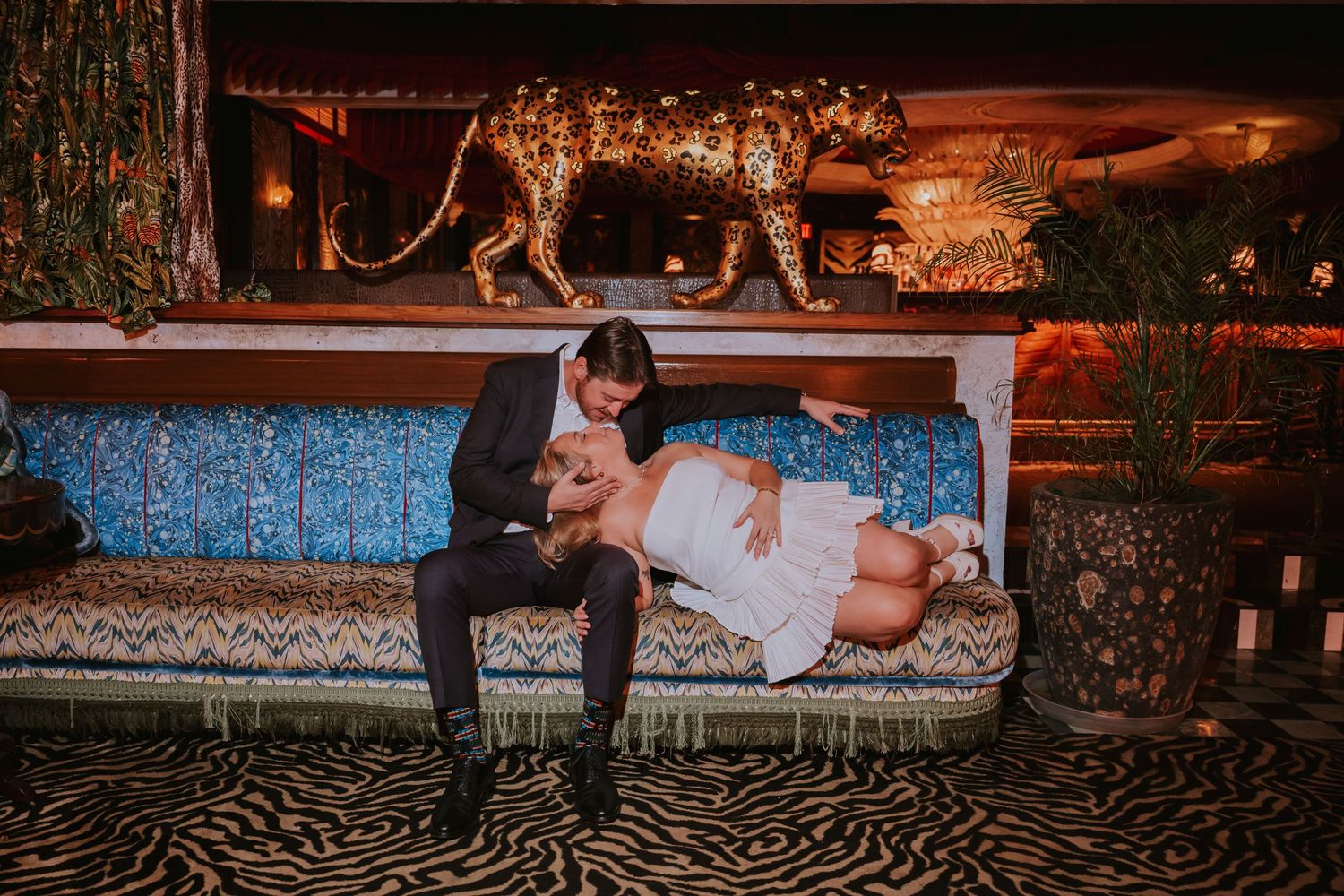 Groom in black suit leans over bride in white dress on a zebra-print sofa beneath a golden leopard sculpture.