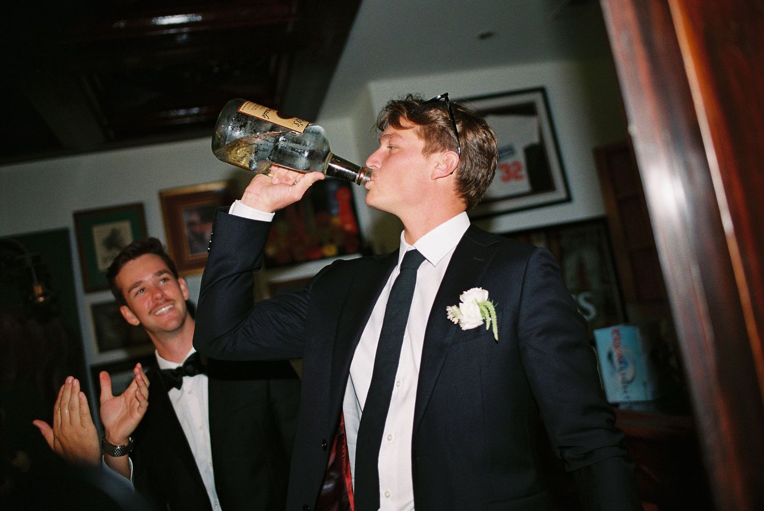 Groom in black suit drinks from champagne bottle at wedding reception while guest in tuxedo applauds.