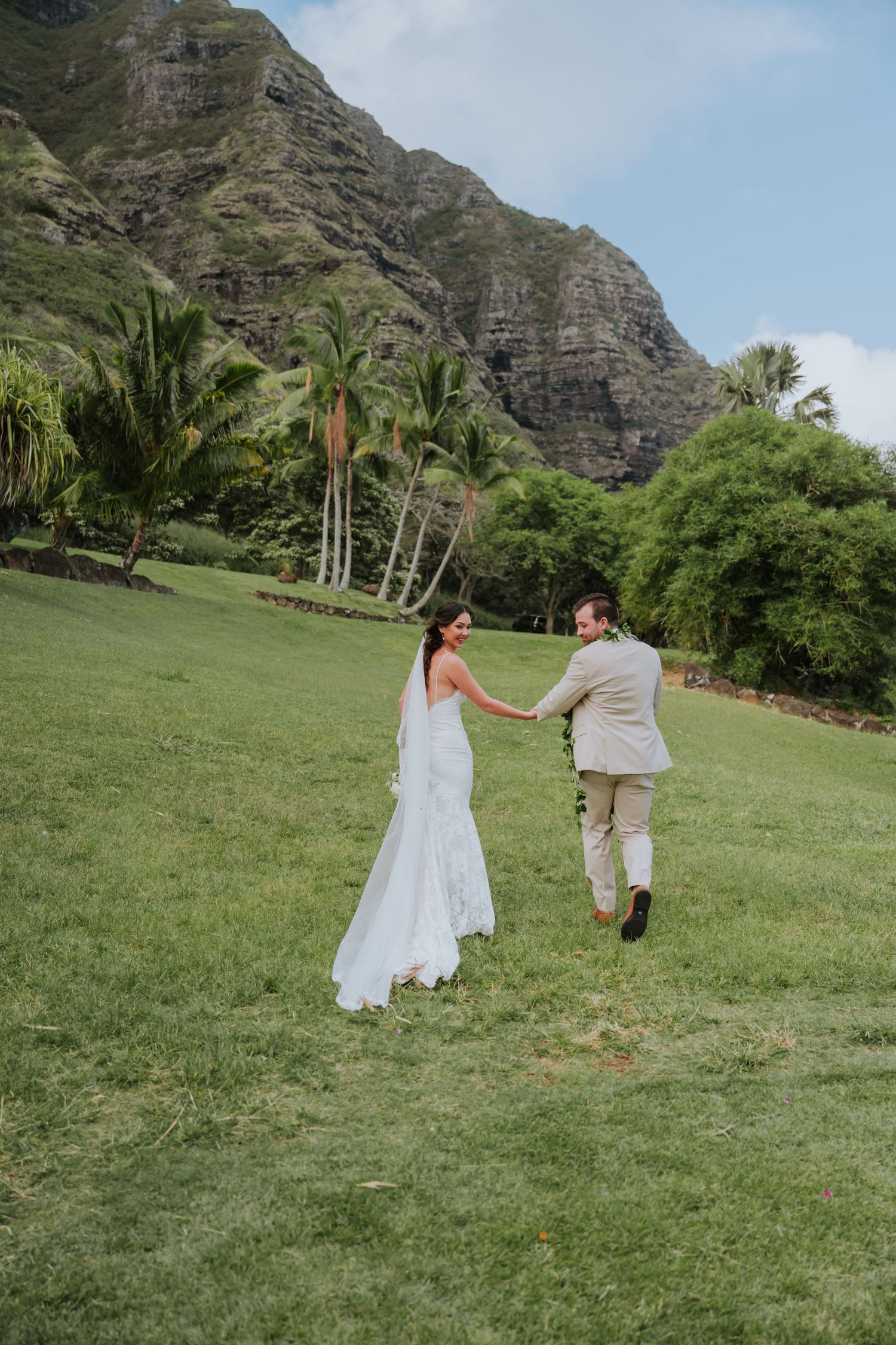 A couple in wedding attire runs playfully across a lush green lawn with dramatic mountain cliffs in the background.