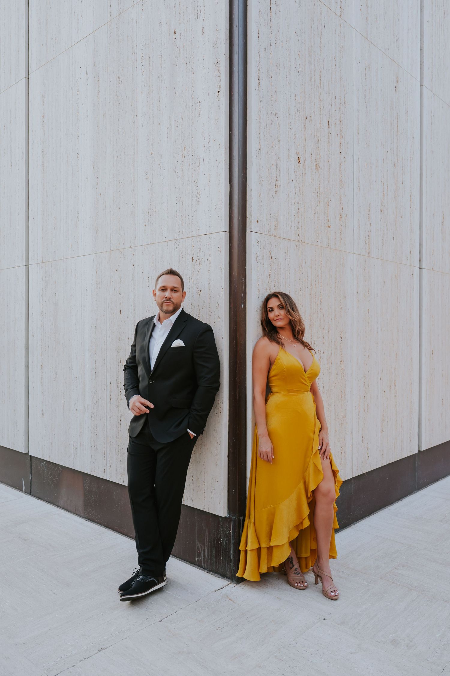 A couple poses against a white wall, with one wearing a black suit and the other in a flowing yellow dress.