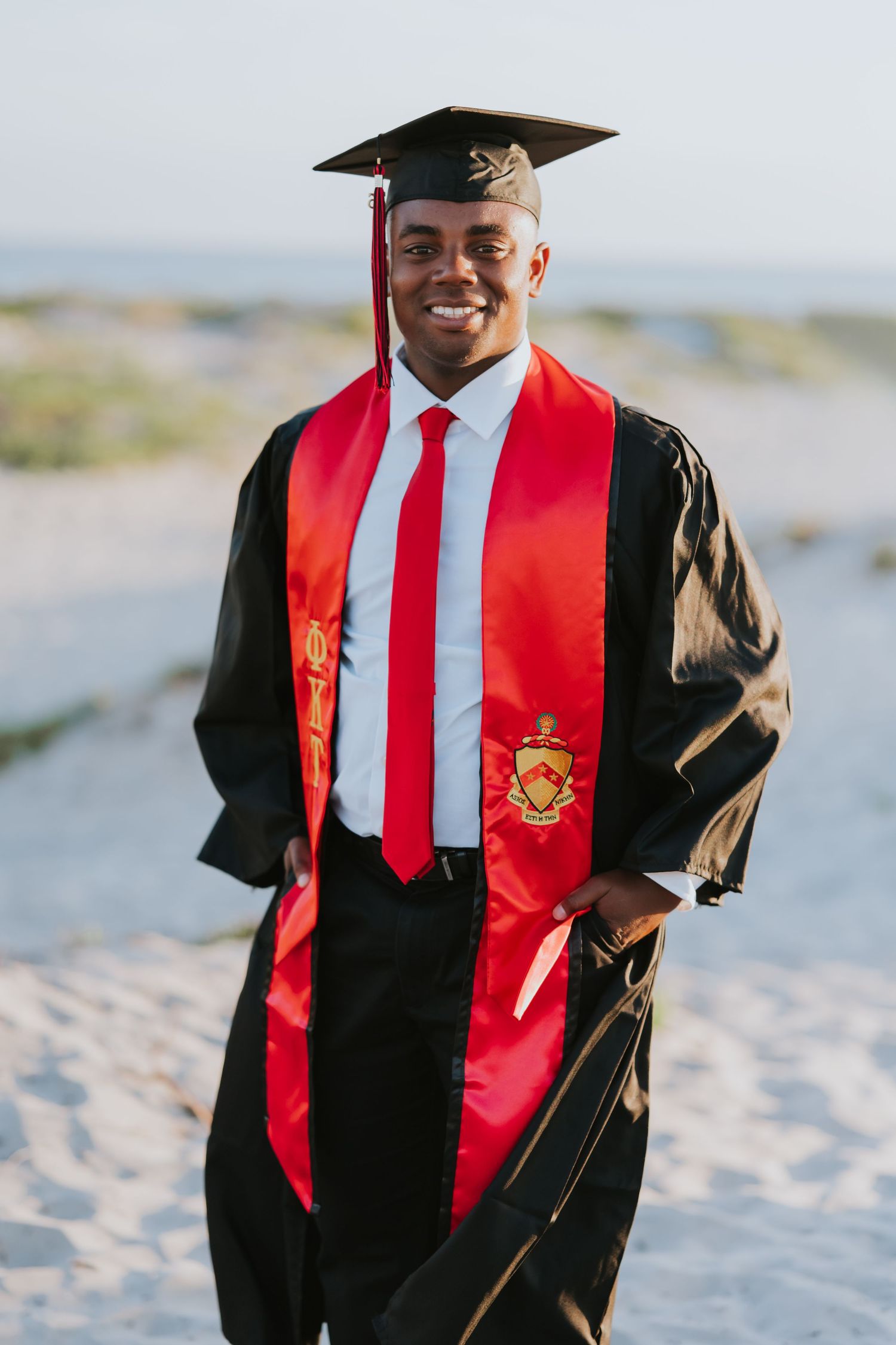 Smiling graduate in black cap and gown with red stole and tie posing on sandy beach at graduation.
