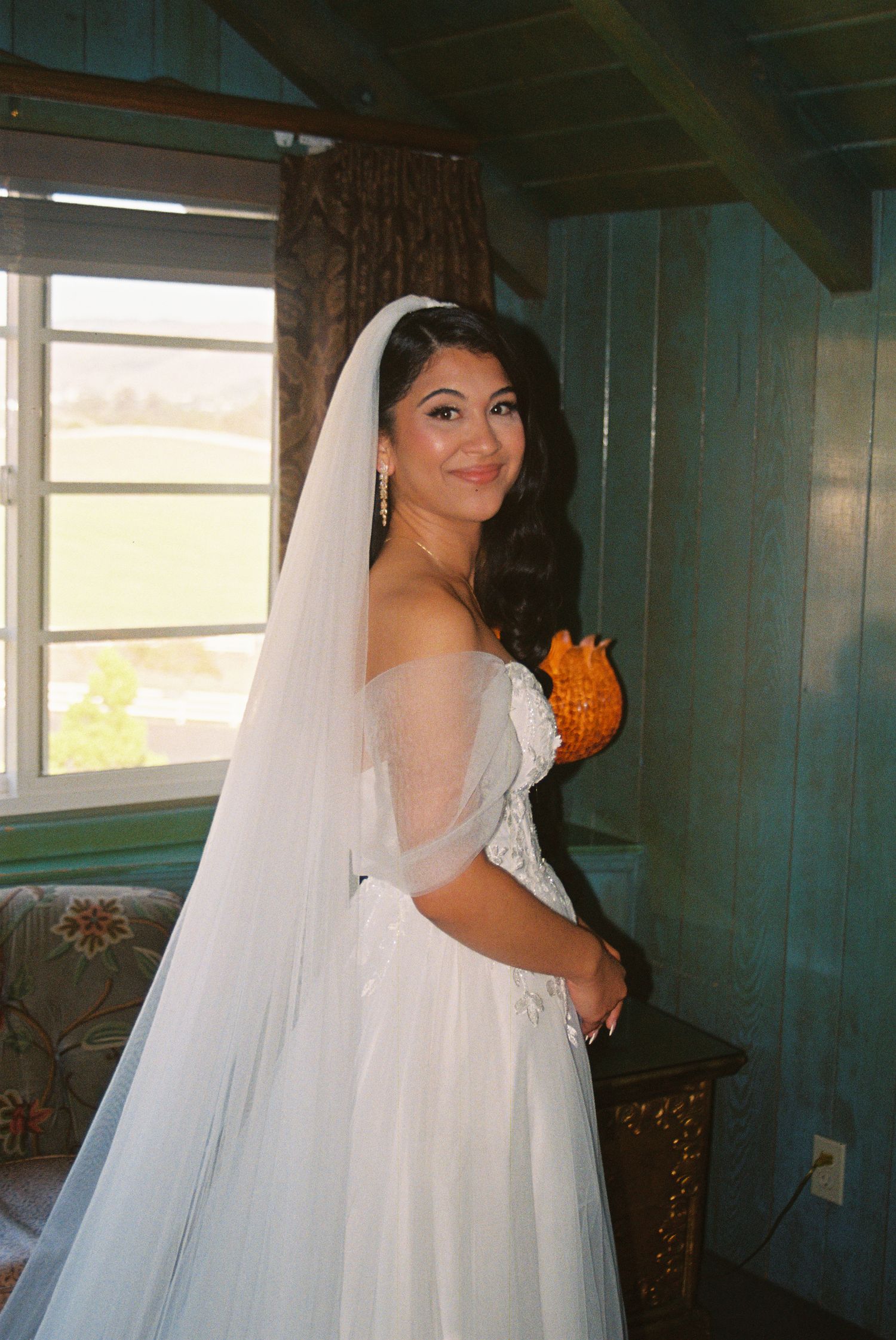 Bride in white strapless gown and veil holding orange bouquet, smiling in rustic blue-walled room.