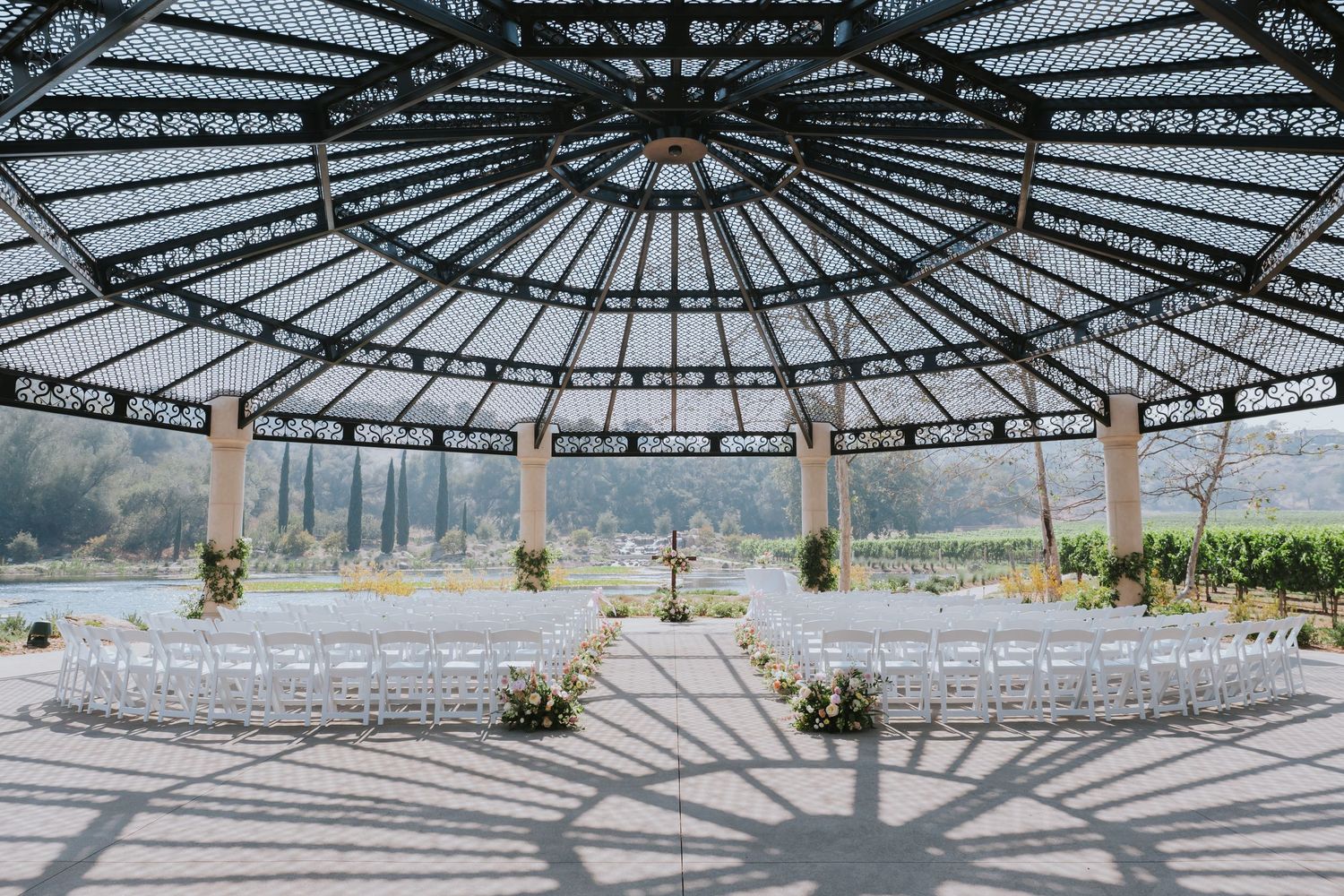 Elegant open-air glass pavilion wedding venue with white chairs arranged for a ceremony, surrounded by scenic vineyard views.