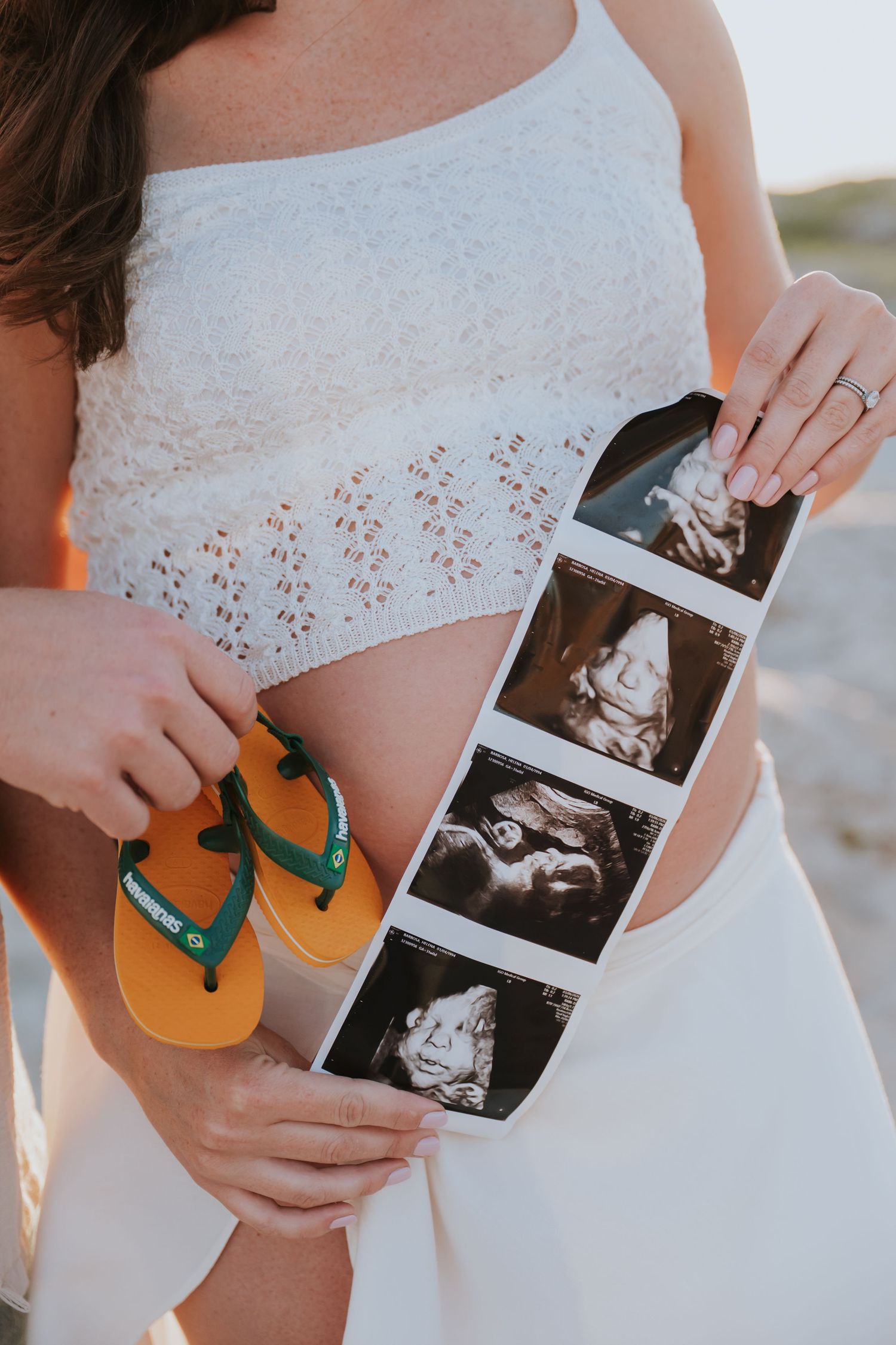 Pregnant woman holding baby ultrasound photos and tiny yellow flip flops against her baby bump outdoors.
