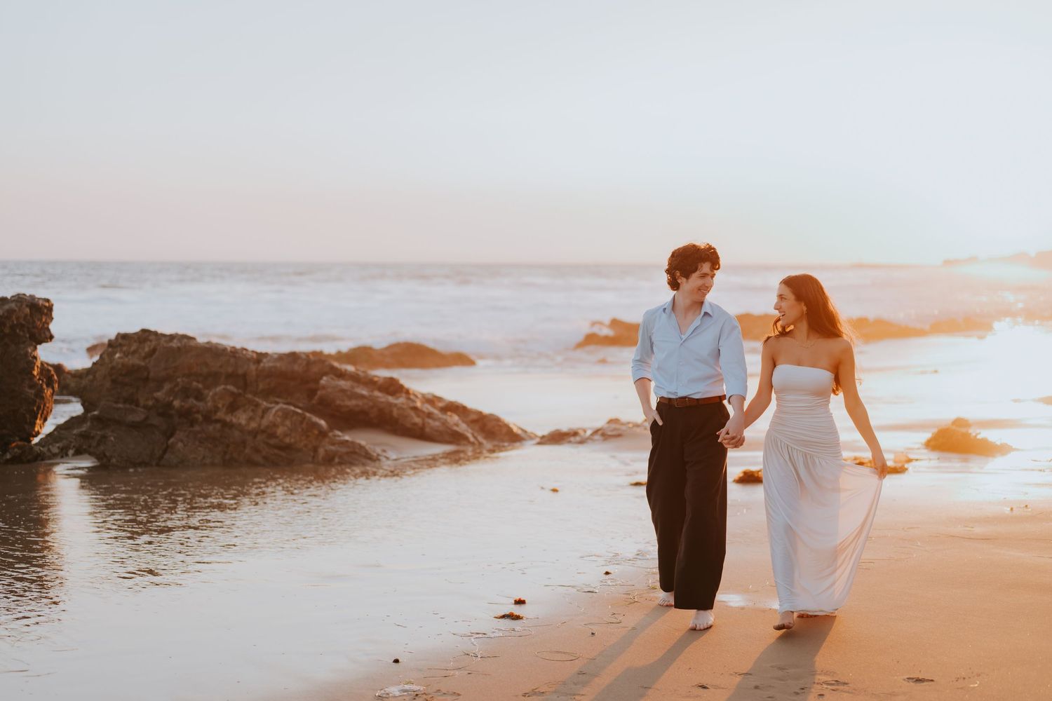 Couple walking hand in hand on a sandy beach at golden hour sunset.
