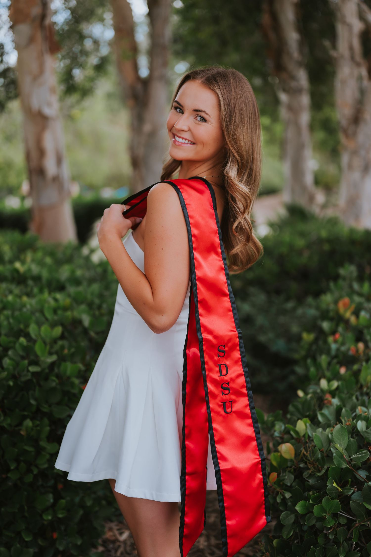Smiling young woman in white dress wearing red SDSU graduation sash poses outdoors among green hedges.