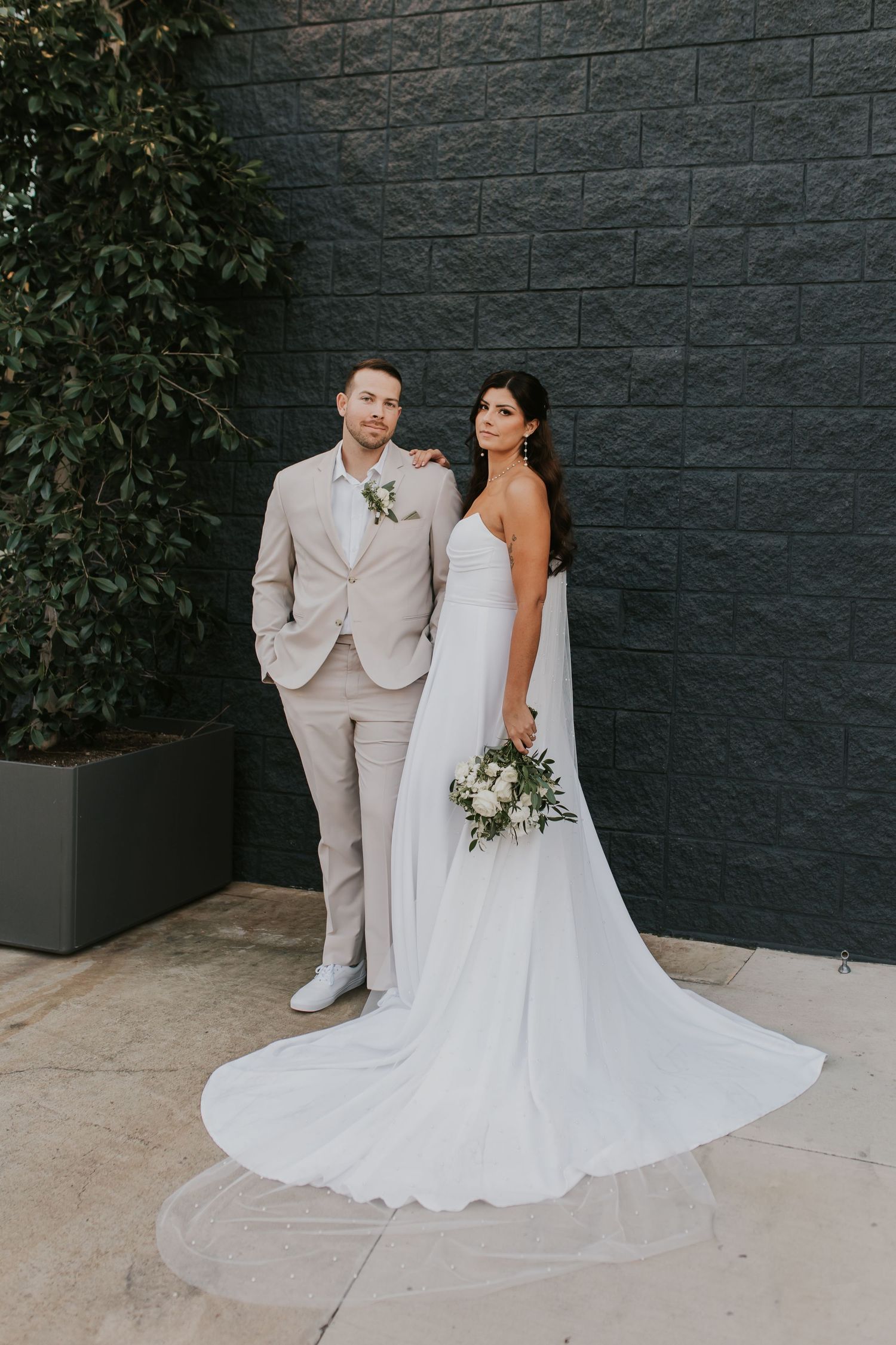 A couple poses against a dark gray wall for their wedding portrait, with the bride in a flowing white gown.