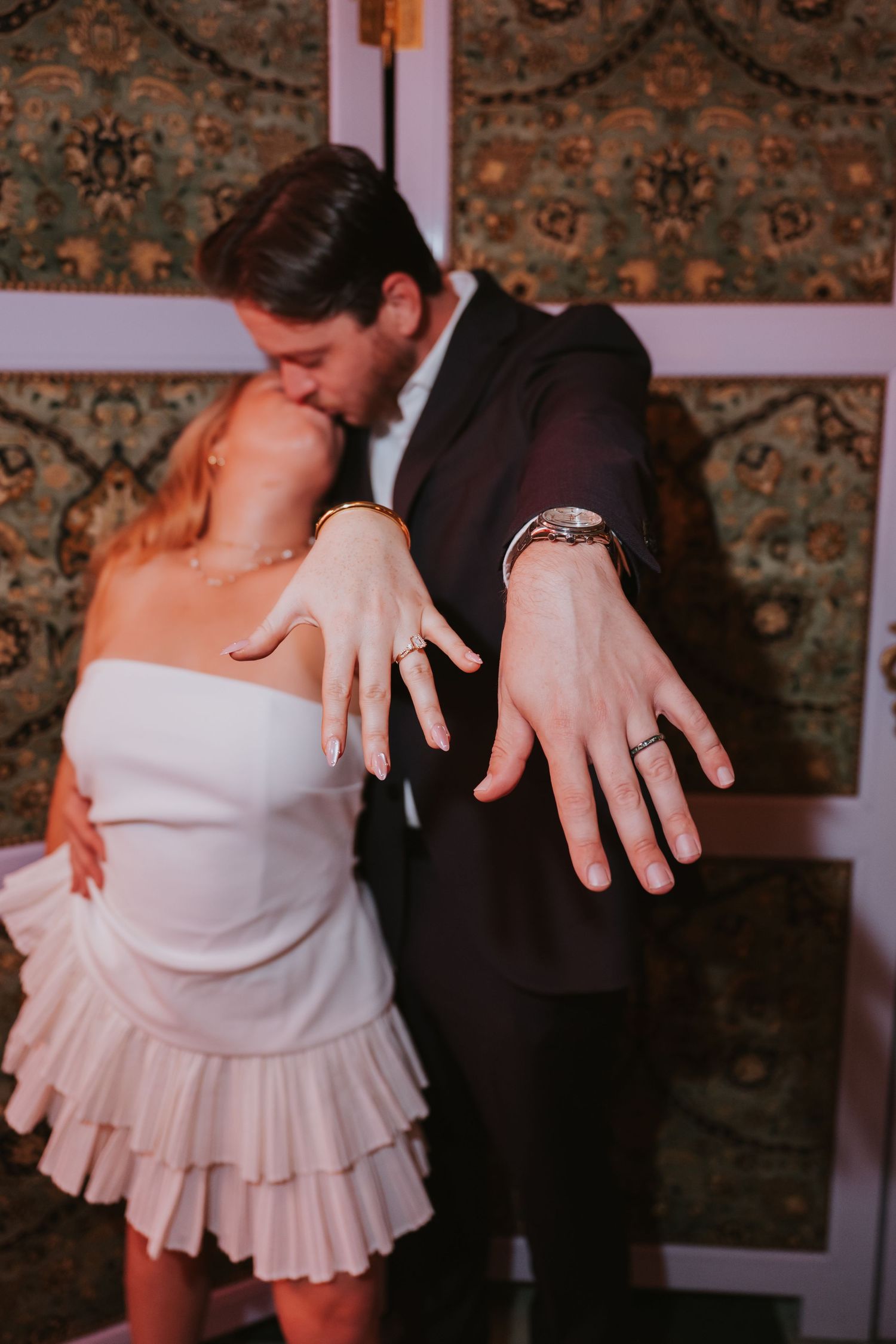 Newlywed couple kissing while showing off their wedding rings to the camera in an elegant decorated room.