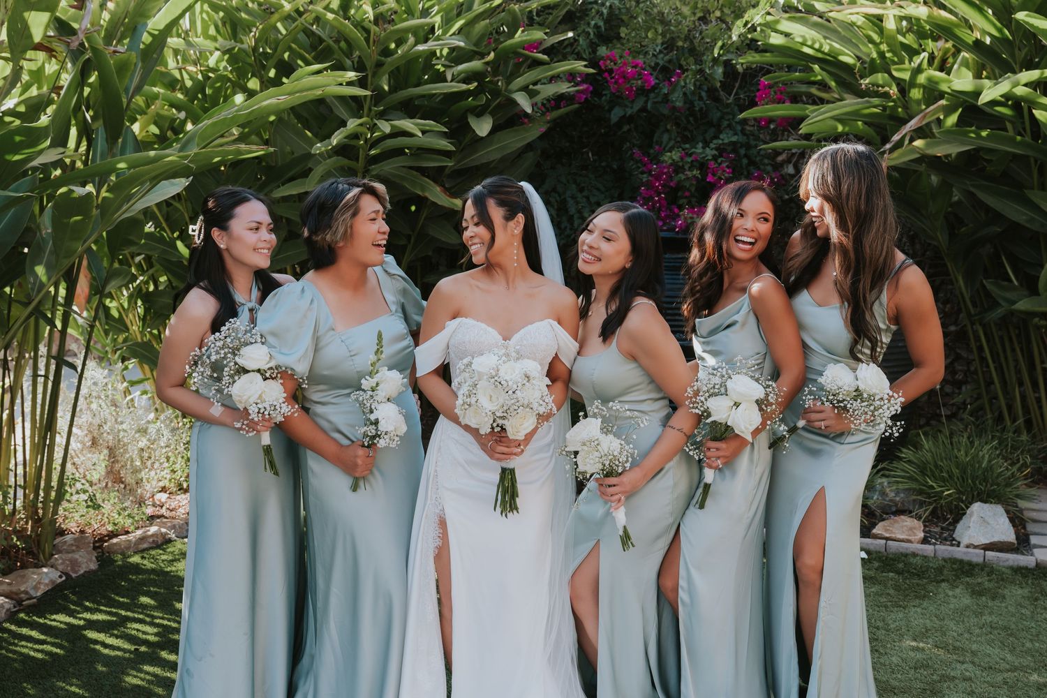 A wedding party in light blue bridesmaid dresses with white bouquets poses together in a tropical garden setting.