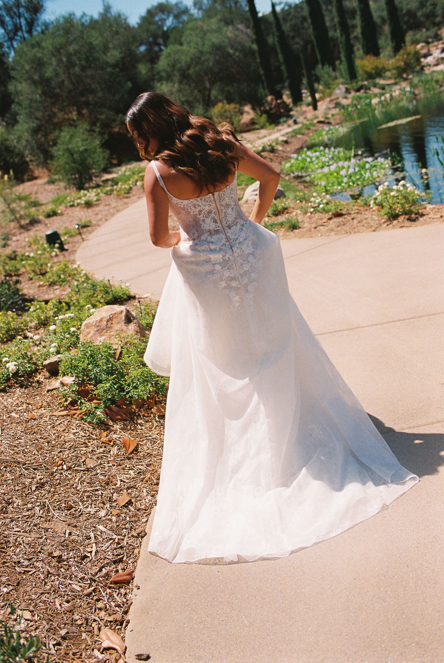 Bride in a white lace wedding gown with a flowing train walks through a garden path.