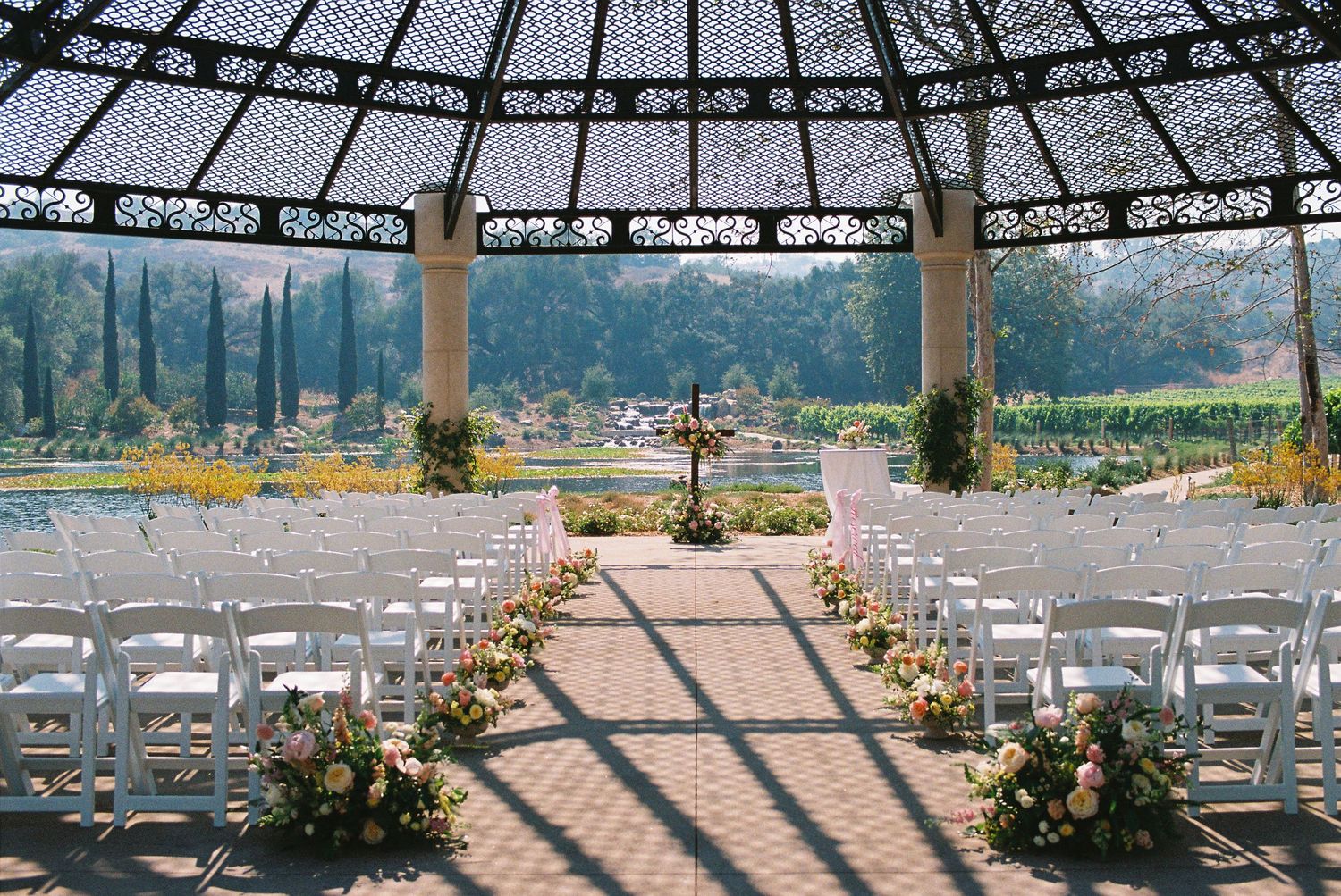 Elegant outdoor wedding ceremony setup under ornate dome gazebo with white chairs, floral arrangements, and lush garden views.