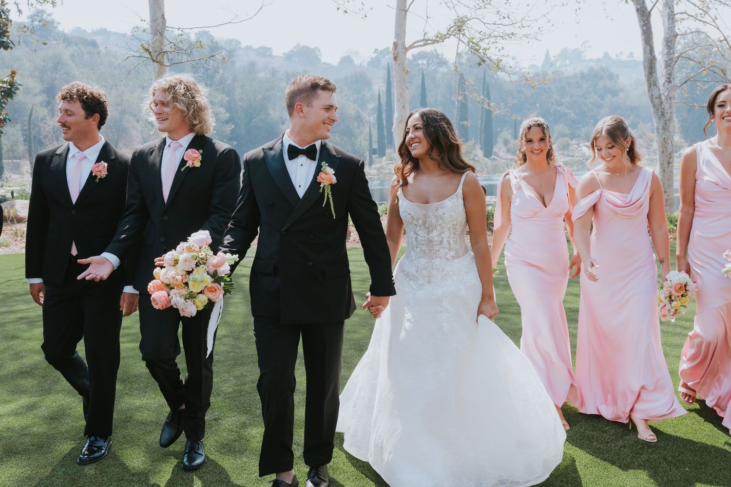 Wedding party walks outdoors, bride in lace gown, groom in tuxedo, bridesmaids in pink dresses, groomsmen in black suits.