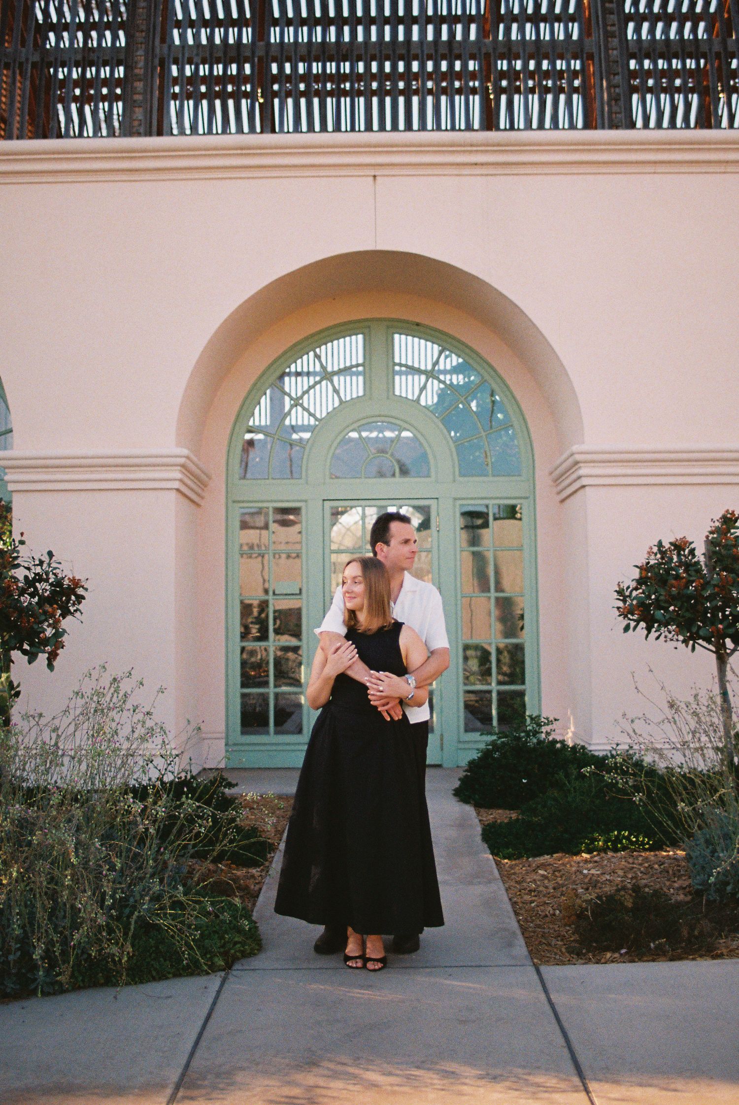 Couple embracing in front of a pink building with arched green door and lush garden landscaping.