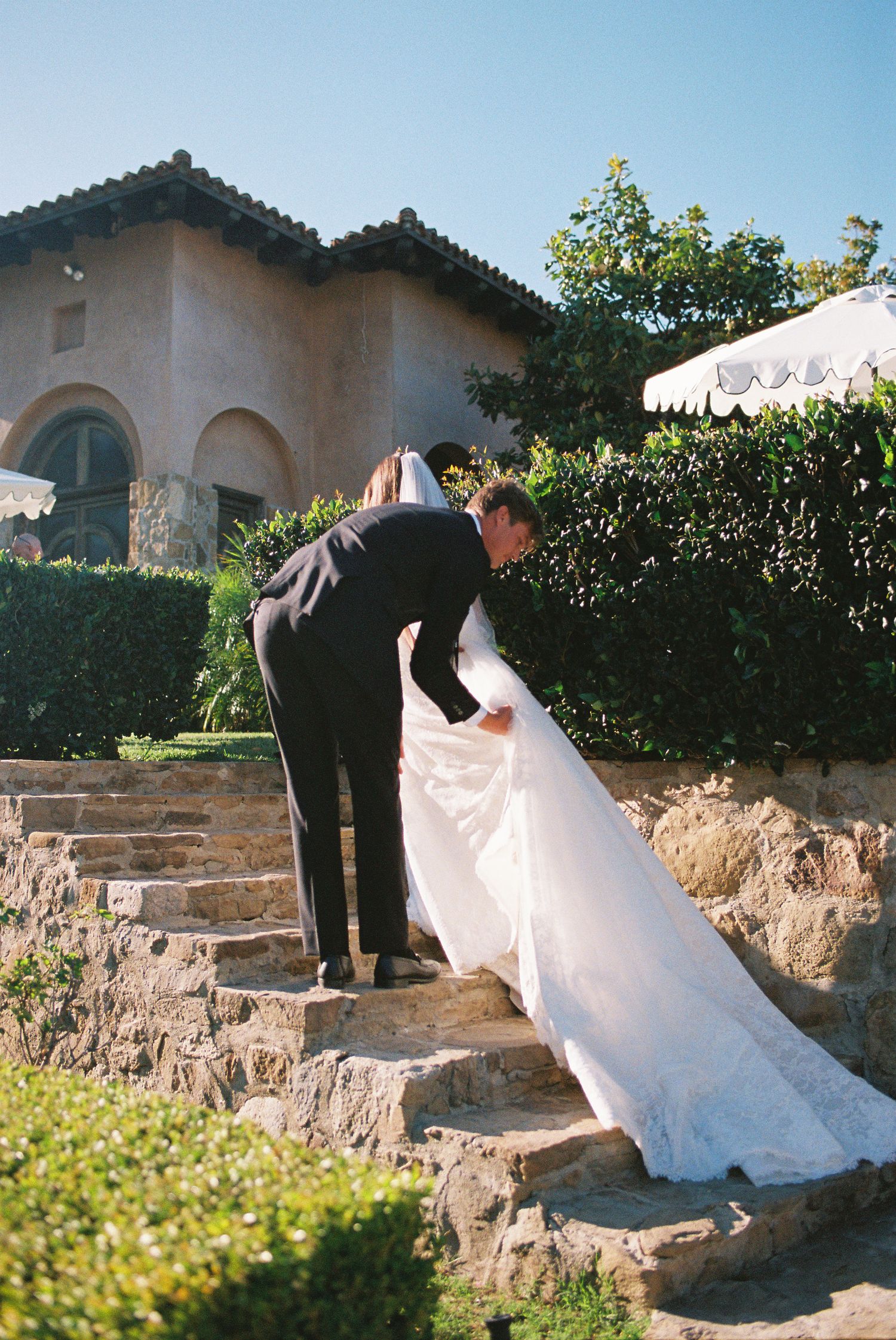 Groom in black suit helps bride in white gown climb stone steps at outdoor wedding venue.