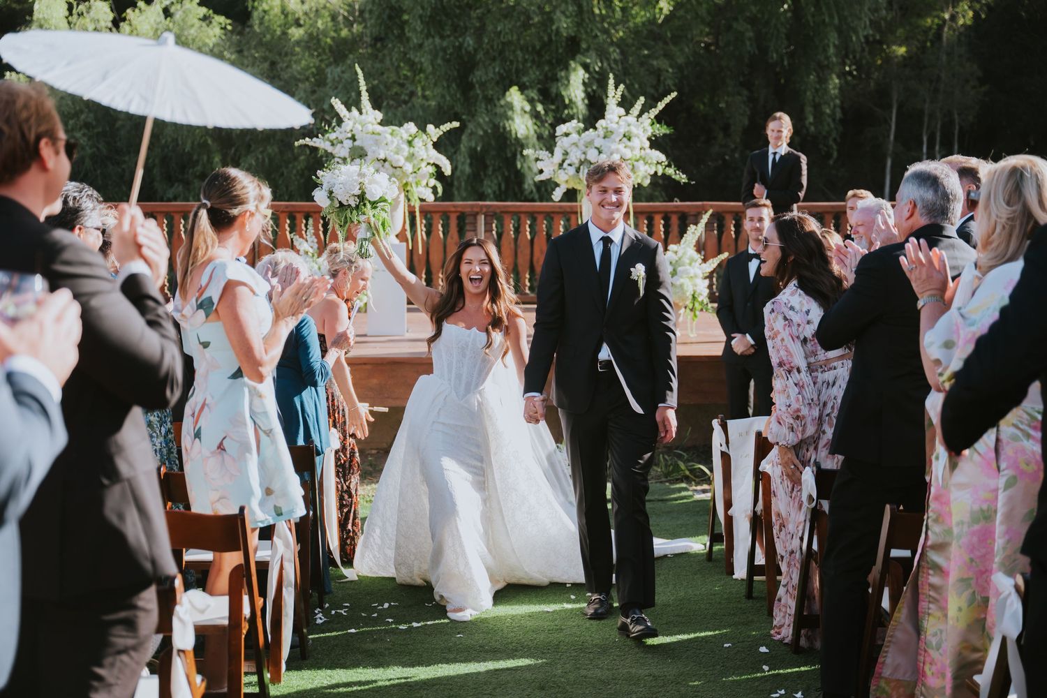 Bride and groom walk down the aisle at an outdoor wedding ceremony surrounded by applauding guests.