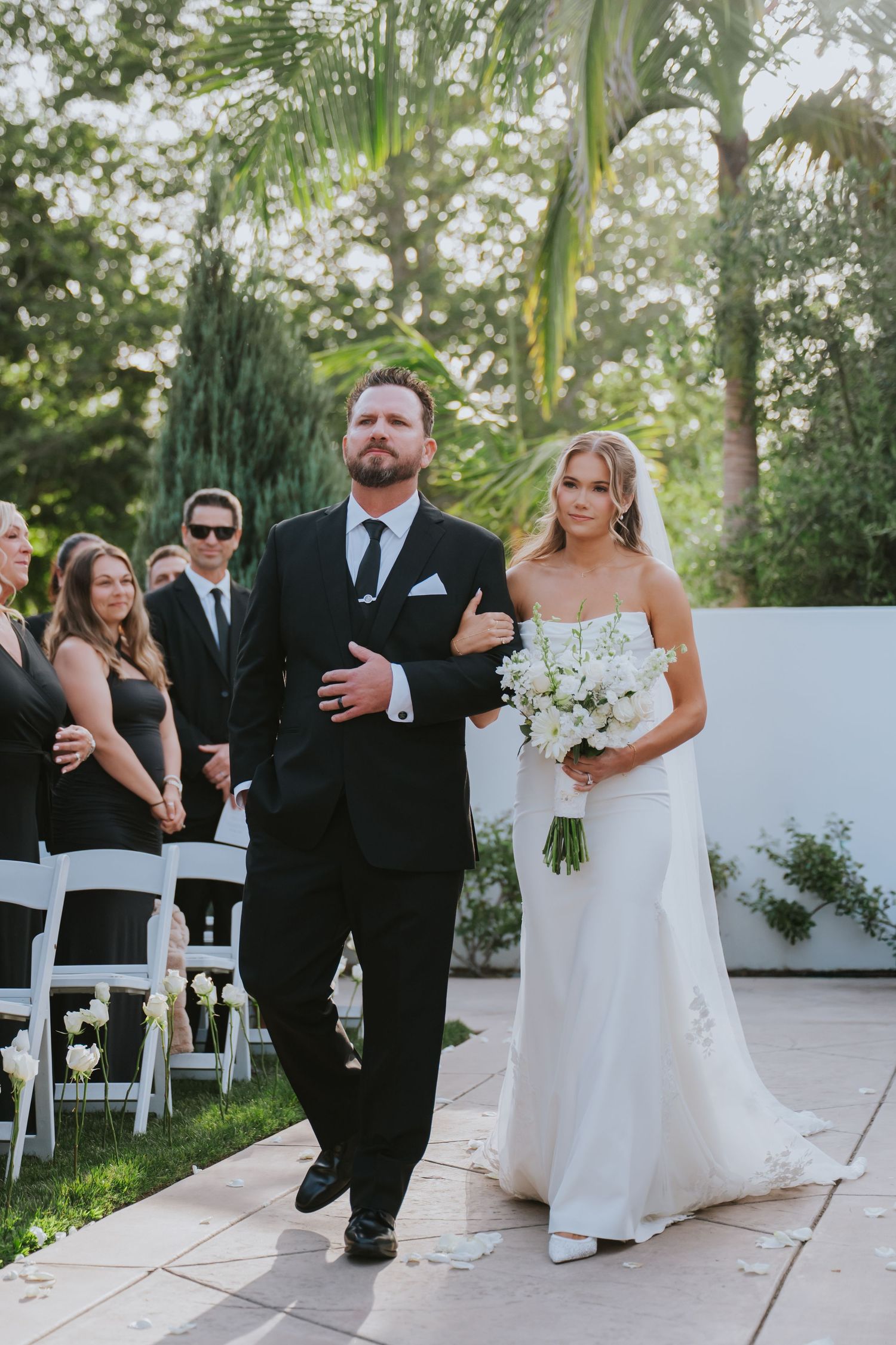 Bride in white strapless gown holding bouquet walks down outdoor aisle with escort in black suit at elegant wedding ceremony.