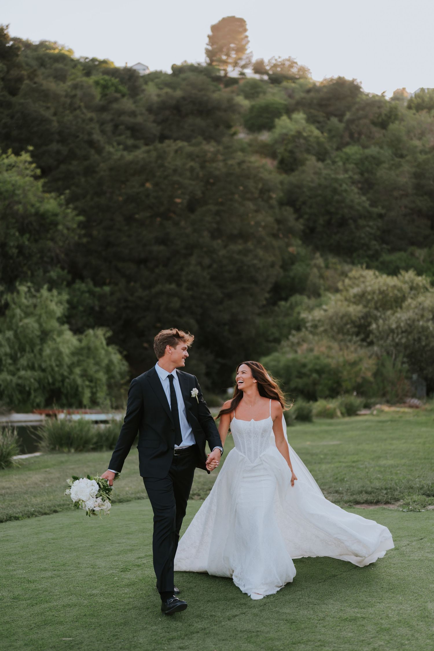 A bride and groom walk hand-in-hand on a green lawn, smiling at each other, with lush hills in the background.