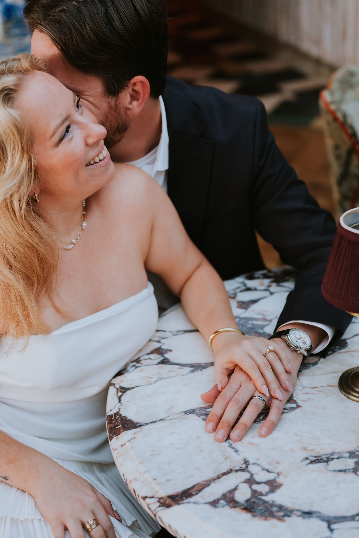 Couple sharing intimate moment at marble table, woman smiling, man in dark suit whispering, holding hands.