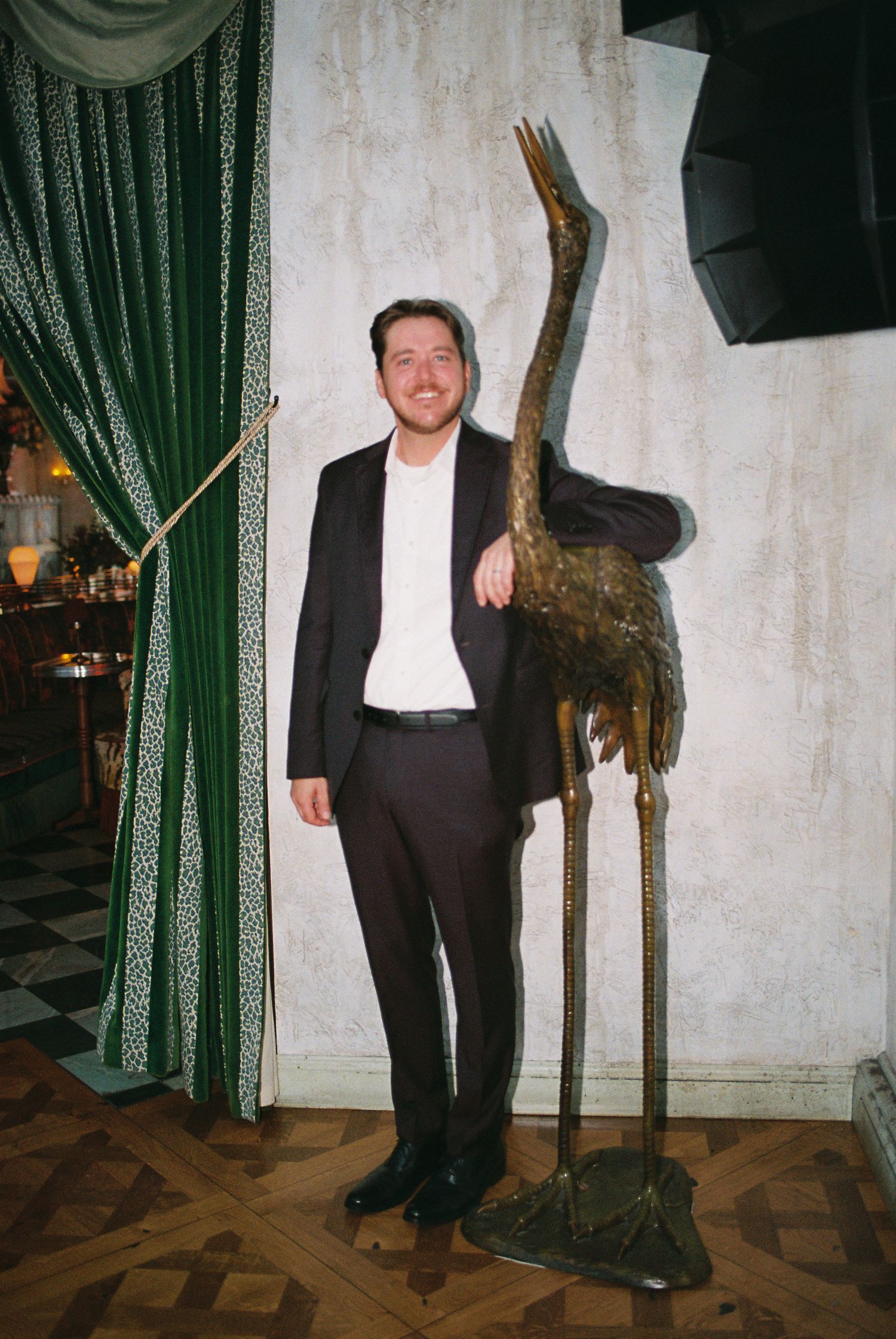 A smiling man in a black suit posing next to a tall bronze crane statue indoors.