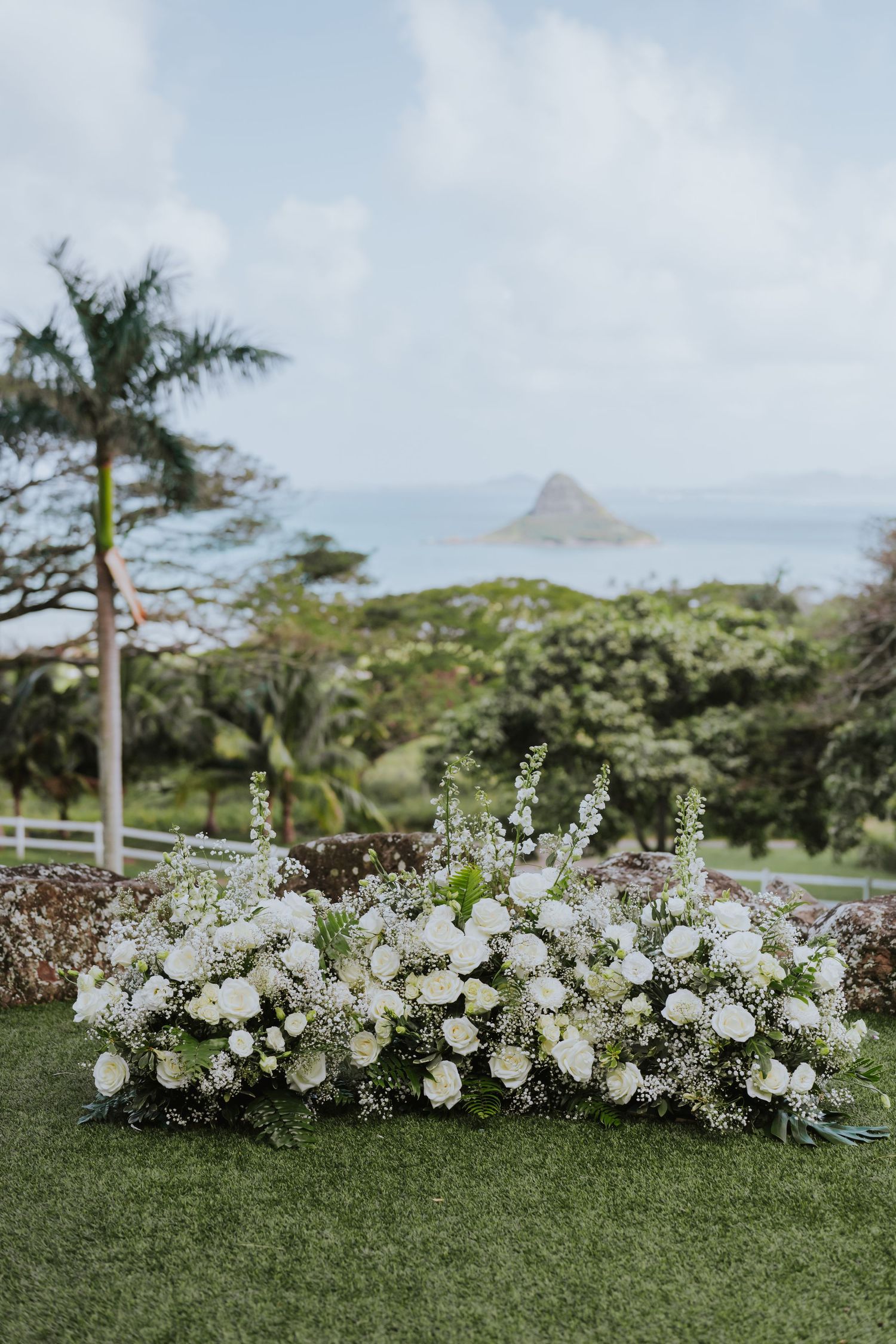 White floral arrangements line a grassy lawn with tropical mountains and palm trees in the background.