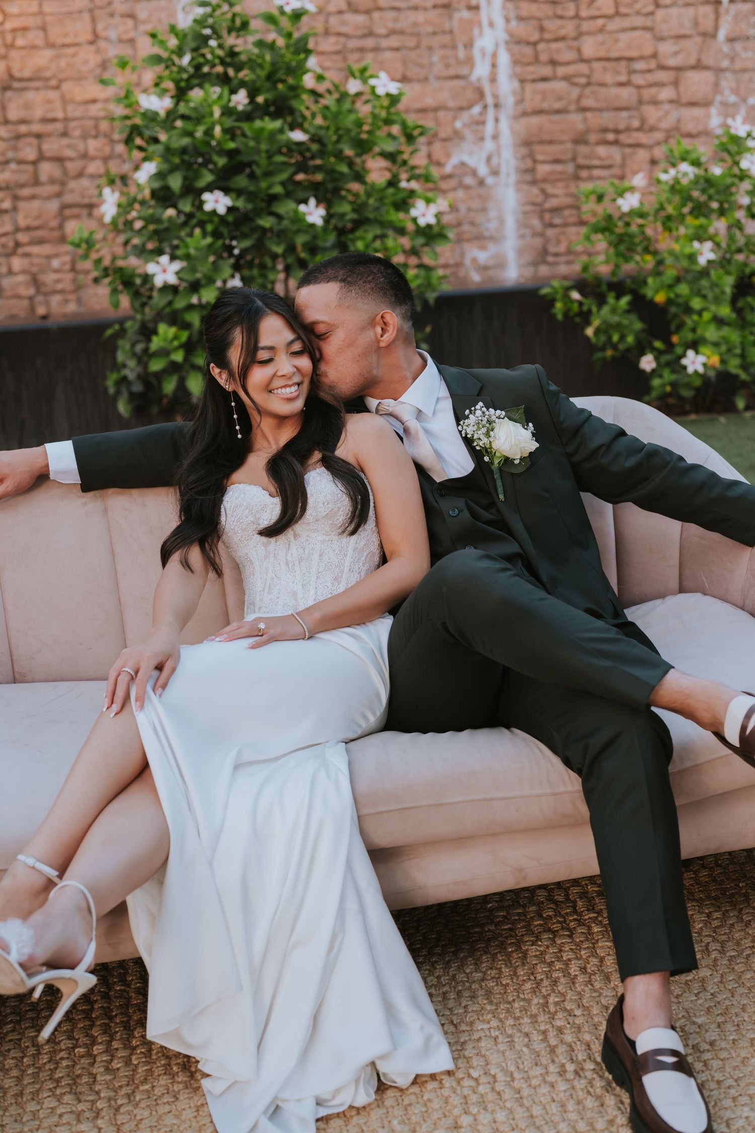A romantic moment between newlyweds on a beige sofa with flowering bushes and a stone wall behind them.