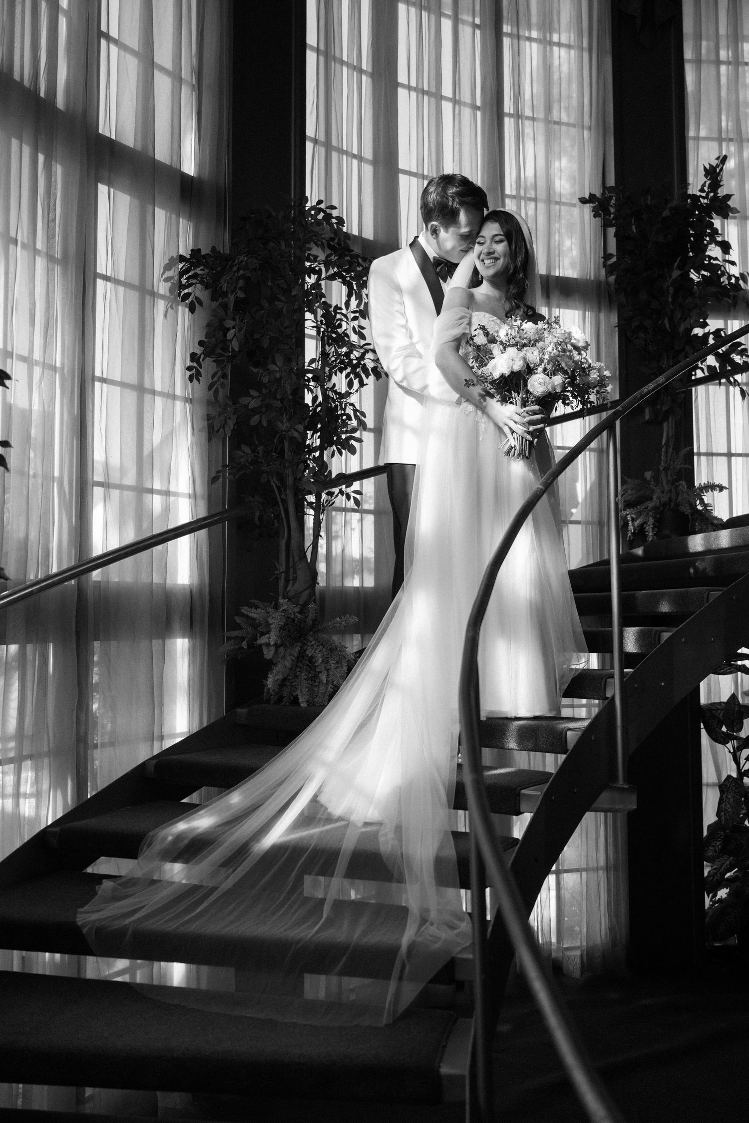 Dramatic black and white photos of a couple in wedding attire by sunlit windows with flowing curtains.