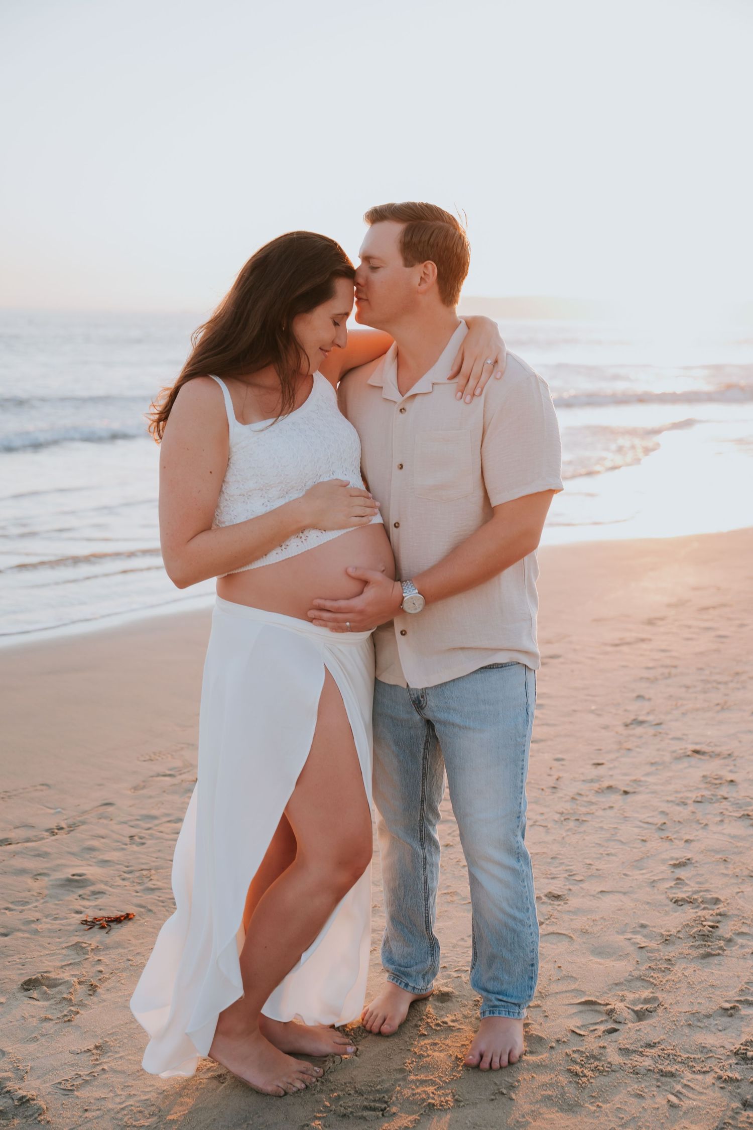 Couple sharing a tender kiss on the beach at sunset, woman in white dress cradling baby bump, man in beige shirt and jeans.