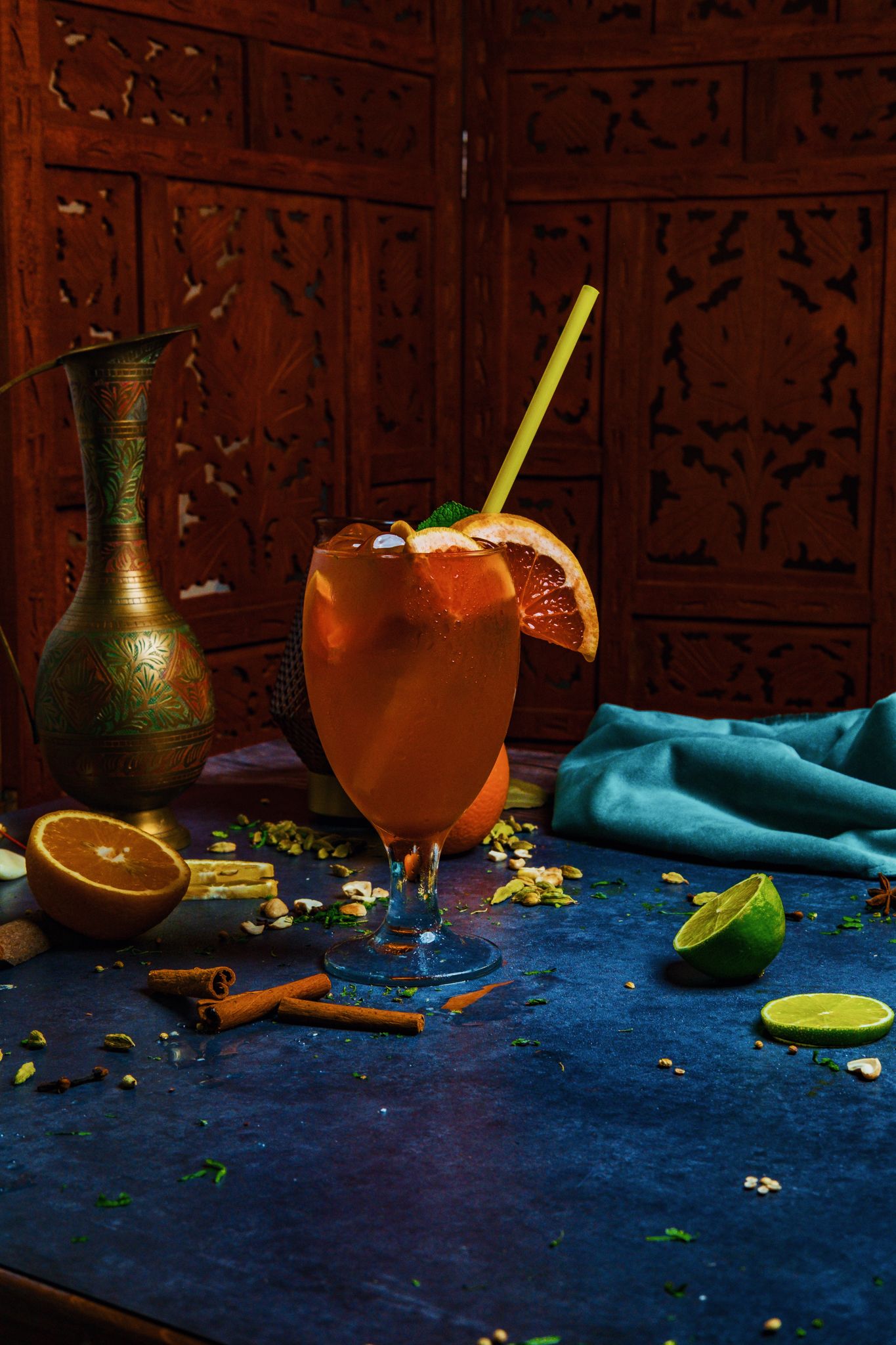 A craft cocktail with citrus garnish served in a wine glass on a blue surface with cinnamon sticks and an ornate backdrop.