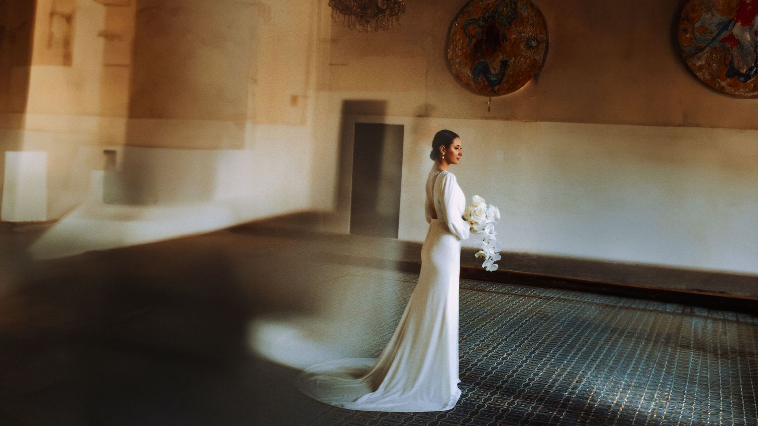 A person in an elegant white wedding gown holds a bouquet while standing in a sunlit hallway with decorative wall details.