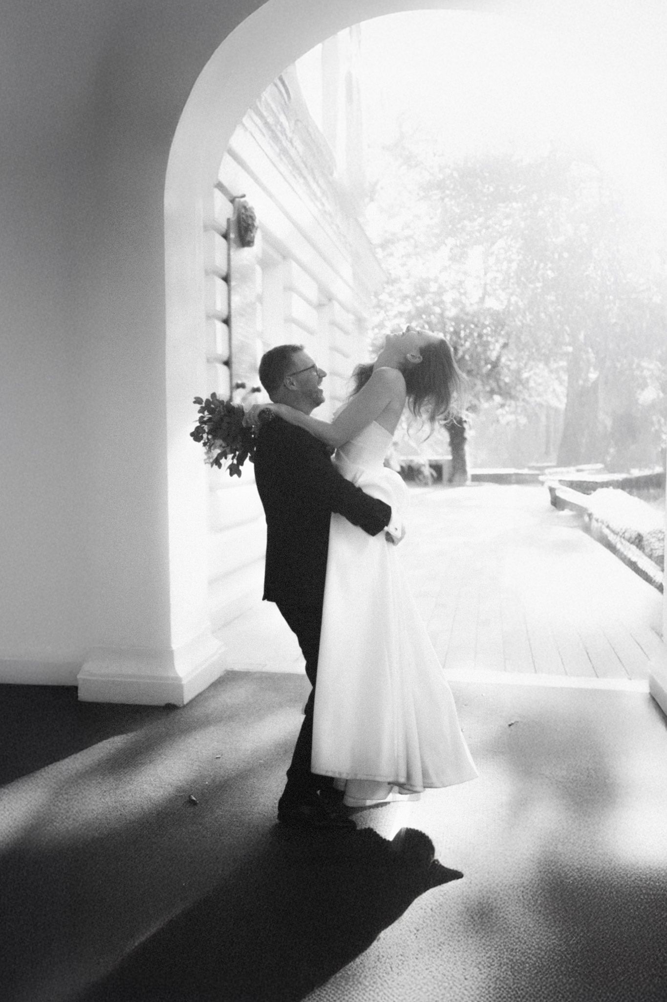 A couple in wedding attire share a romantic dance together under an arched doorway in black and white photography.