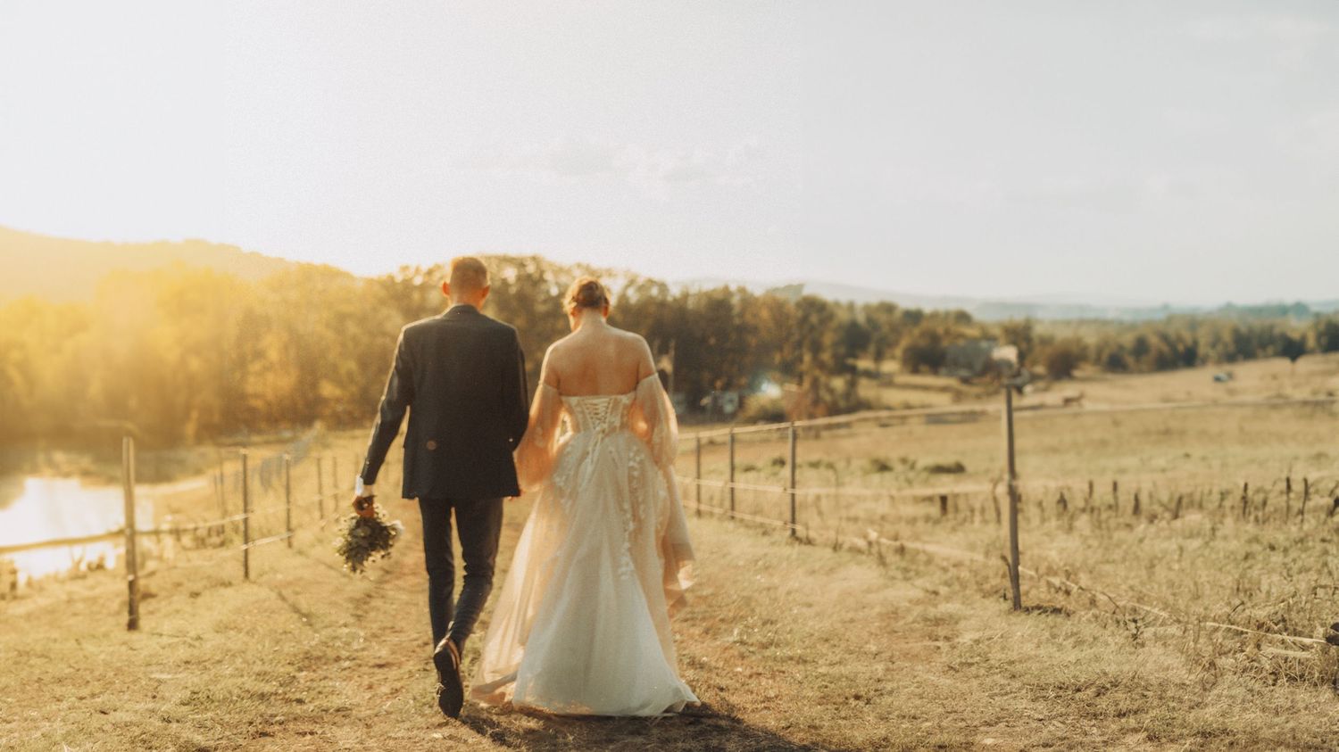 A couple walks hand in hand along a rural path at sunset, with the sun casting a warm glow over the countryside.