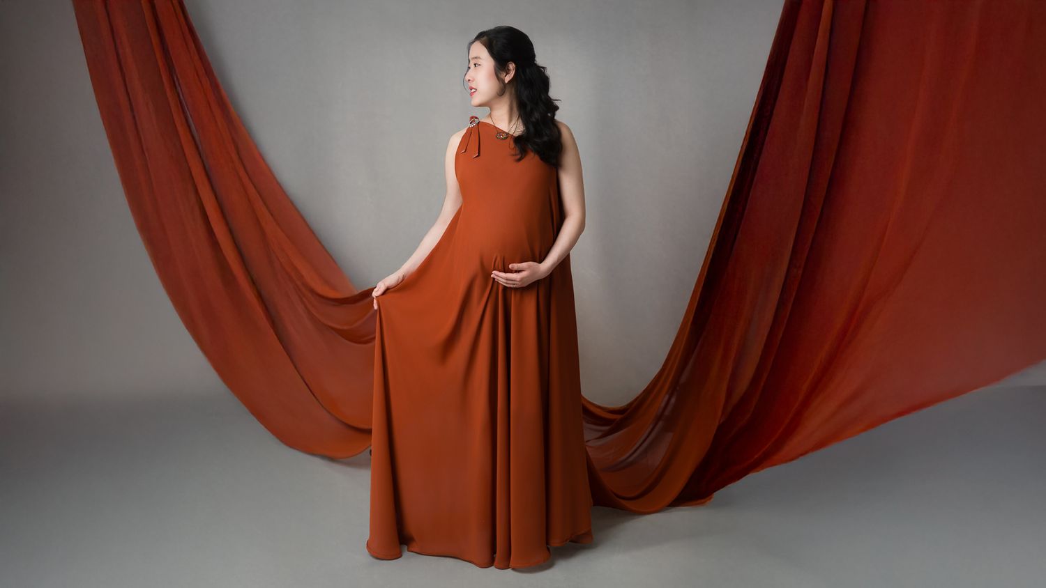 Maternity portrait of a pregnant woman in a chiffon gown, gently cradling her baby bump. Captured in a drastic draps, side-profile