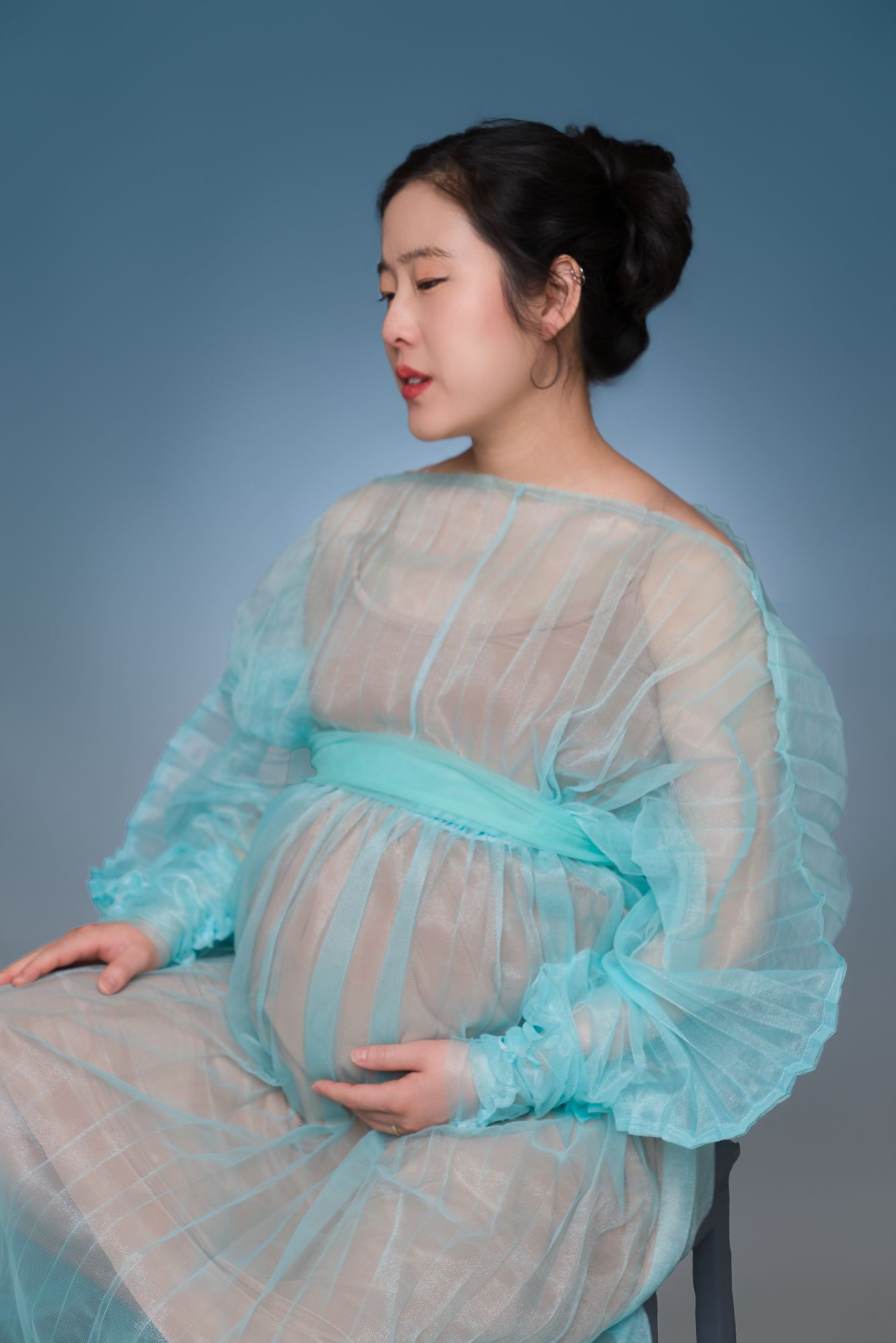 Maternity portrait of a pregnant woman in a flowing sheer turquoise gown, gently cradling her baby bump. Captured in a serene, side-profile