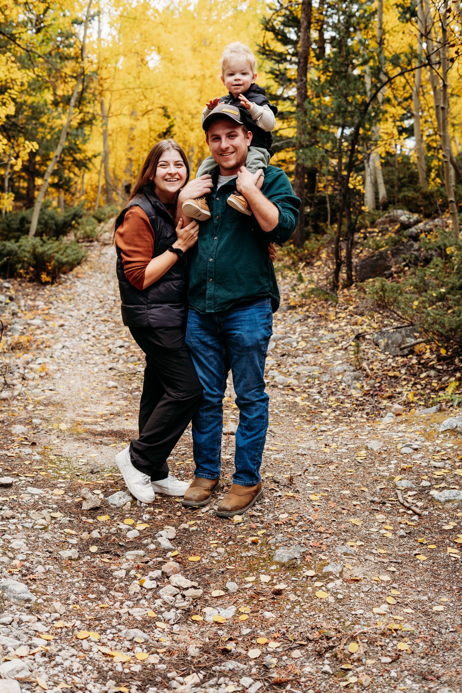 Candid outdoor family photography session featuring parents and their toddler son smiling at each other among yellow fall leaves in Colorado