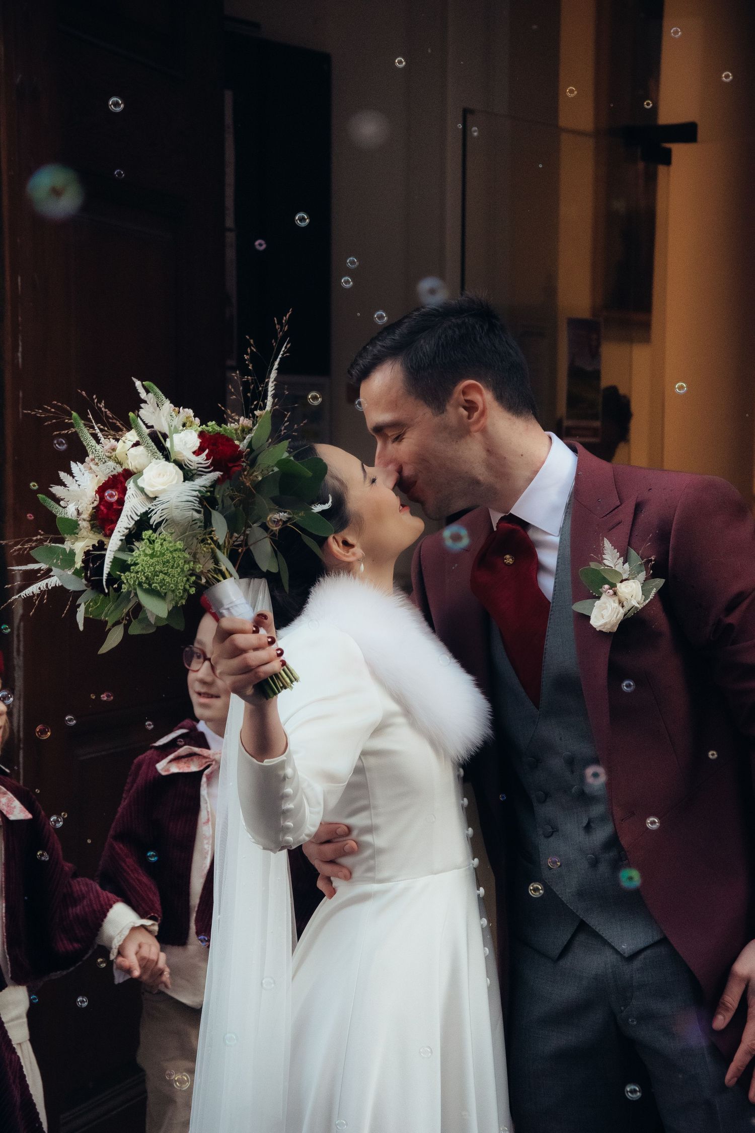 Newlyweds share romantic moment with white bouquet and burgundy suit.