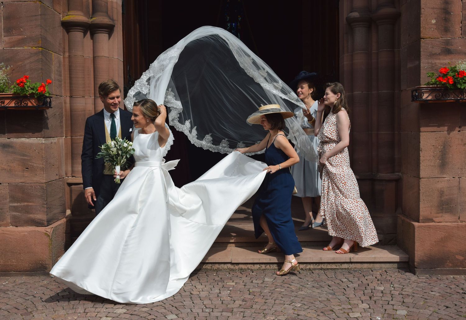 Wedding party helps adjust bride's flowing veil in front of stone church entrance.
