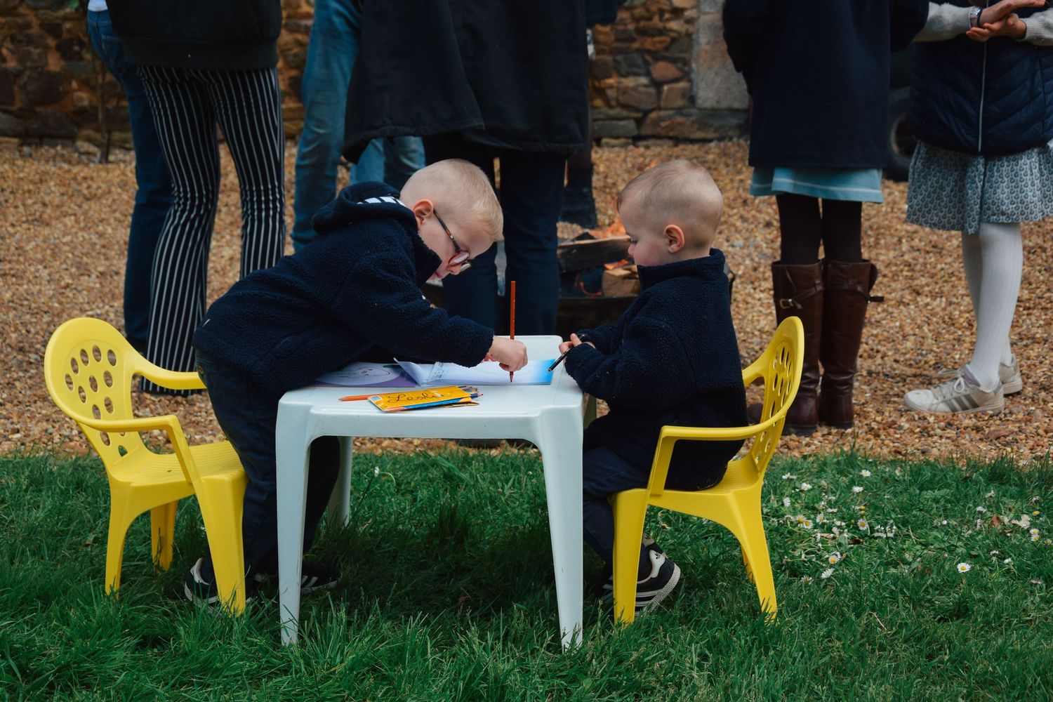 Children sitting at a small white table with yellow chairs outdoors.