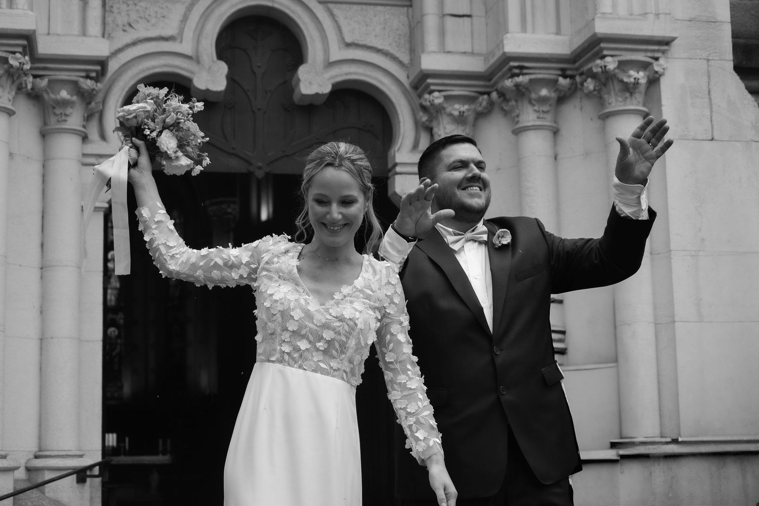 Newlyweds celebrate joyfully with raised arms outside an ornate church entrance in black and white photograph.
