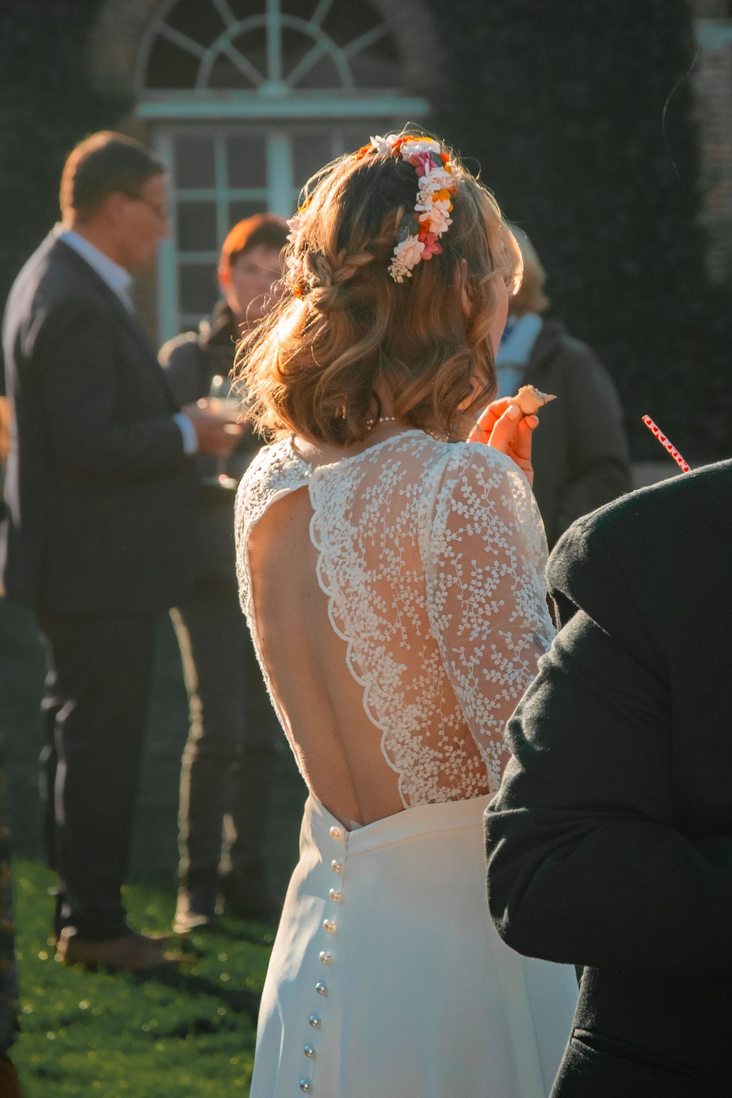 Back view of a bride in an elegant lace-back wedding dress with floral crown during an outdoor garden ceremony.