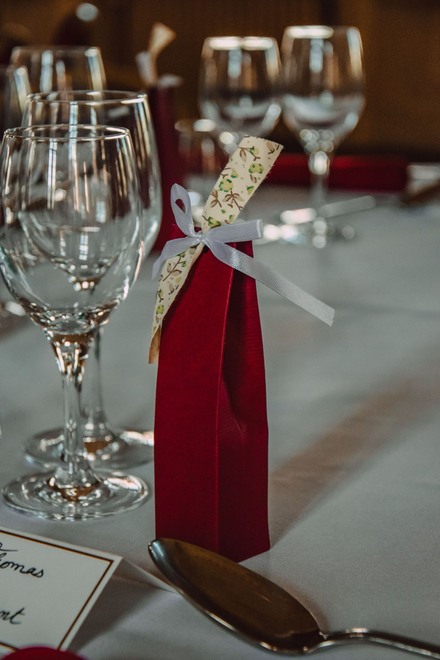 Wedding place setting with wine glasses and red favor bag tied with decorative ribbon.