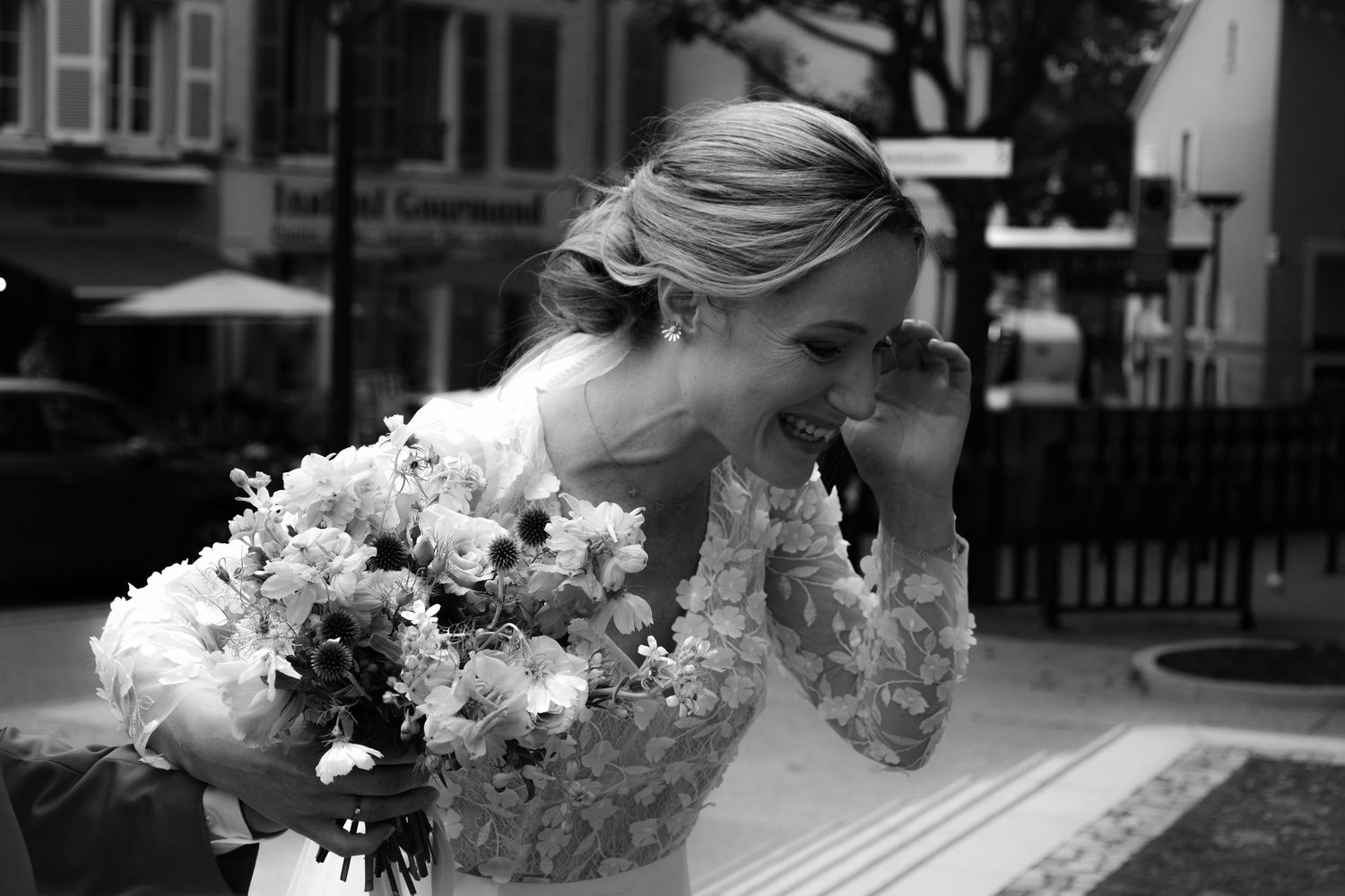 Black and white wedding photo showing bride in lace dress holding a white floral bouquet while looking down.