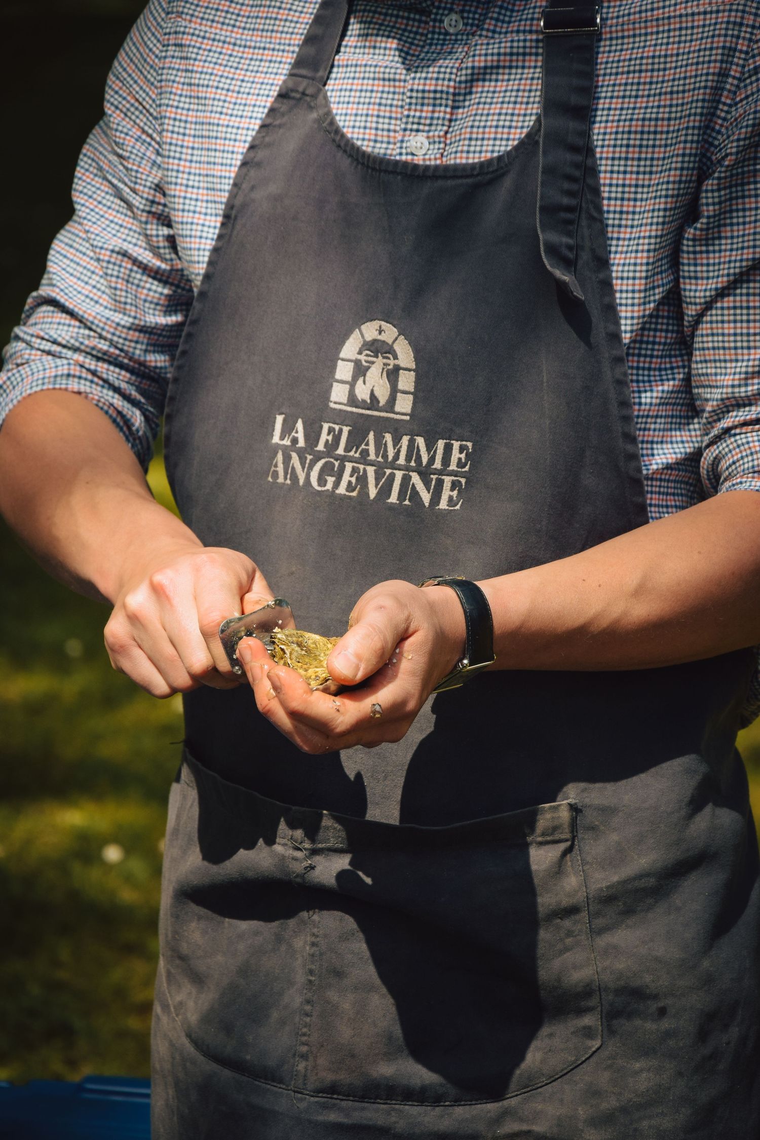 Chef wearing La Flamme Angevine apron prepares ingredients in sunlight.